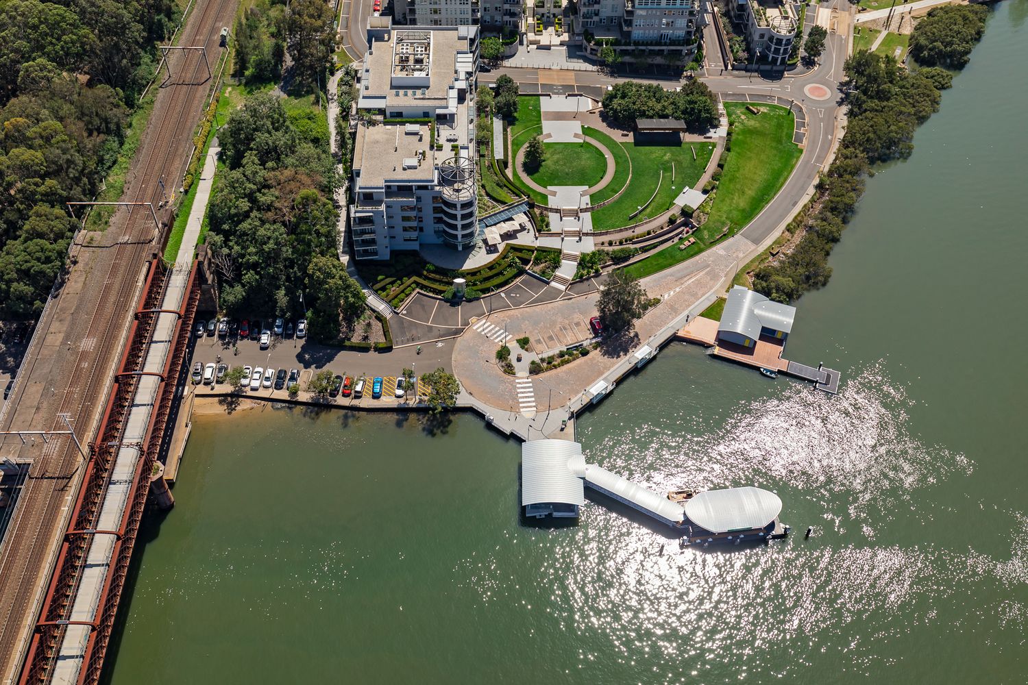 Aerial view of a curved ferry wharf with walkway connecting to waterfront apartments, parkland and a nearby railway line.