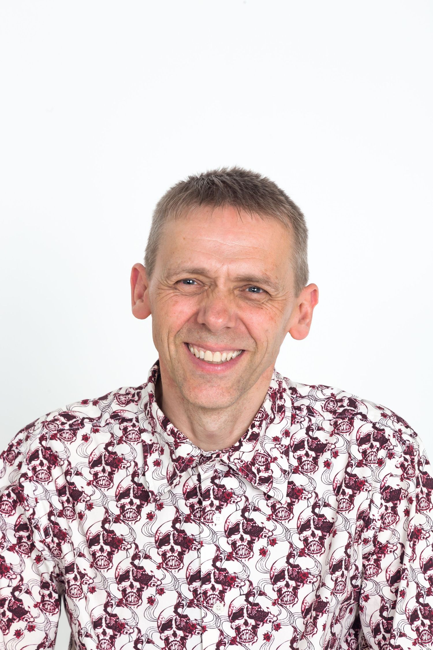 A smiling middle-aged man with short gray hair wears a patterned skull print shirt against a clean white studio background.