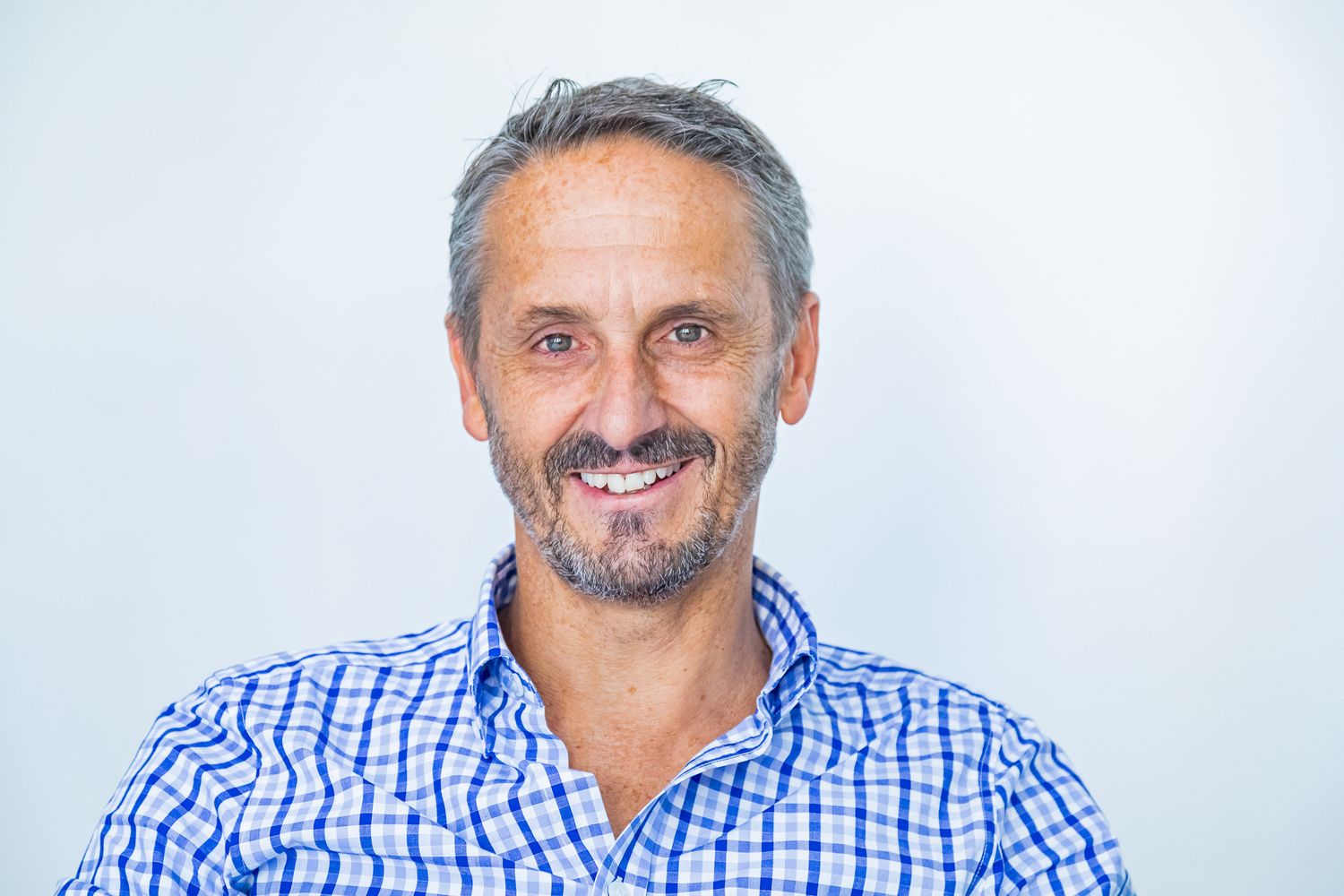 Smiling middle-aged man with graying hair and a salt-and-pepper beard wearing a blue checkered shirt against a bright white background.
