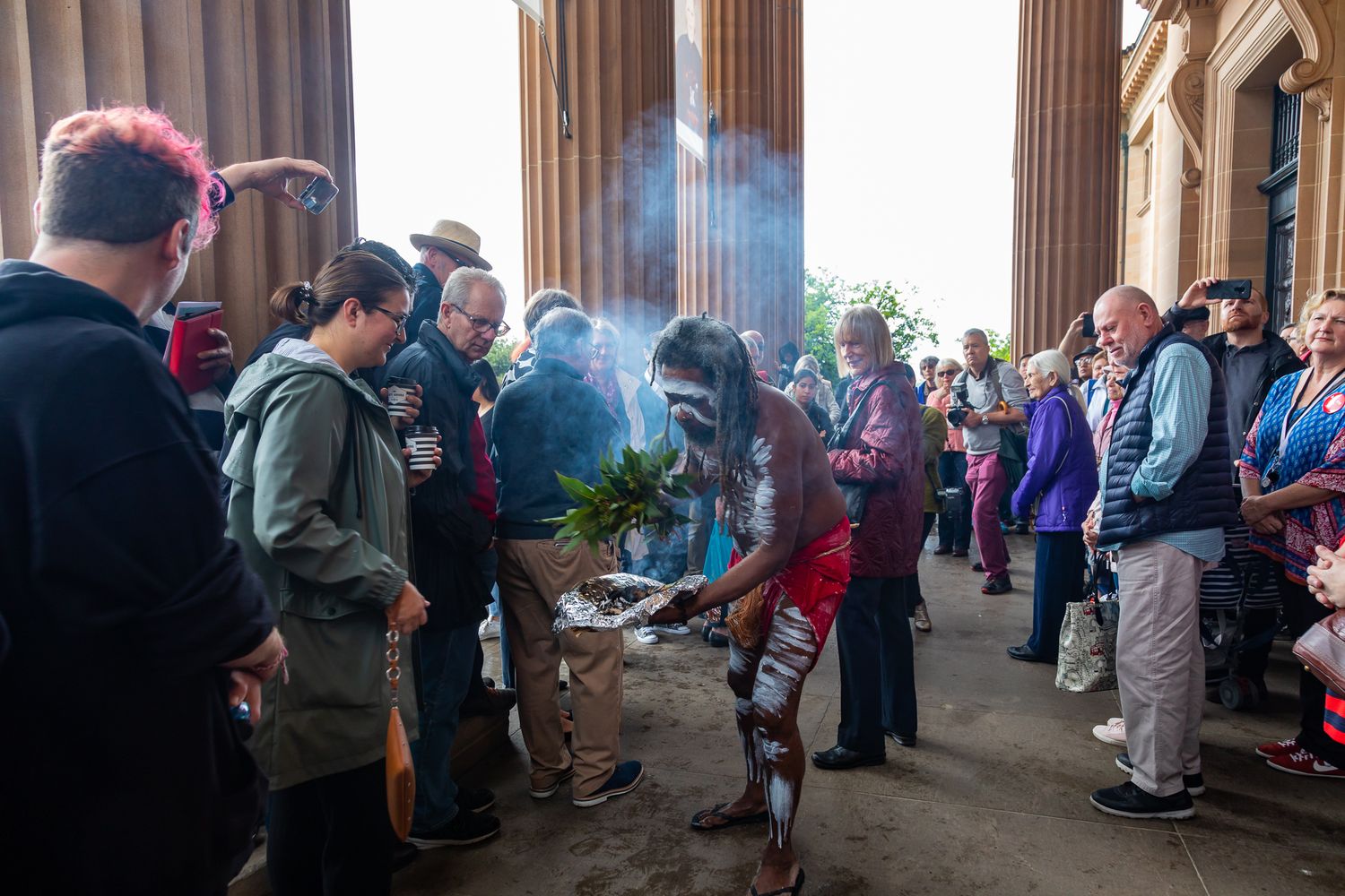 Performer with body paint holds smoking native leaves in a smoking ceremony as a crowd gathers outside a columned building.