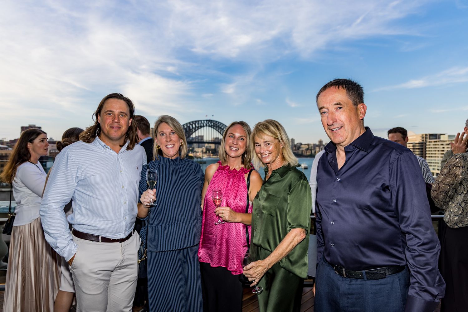 Group of five smiling guests hold drinks at a waterfront event with the Sydney Harbour Bridge visible under a blue sky.