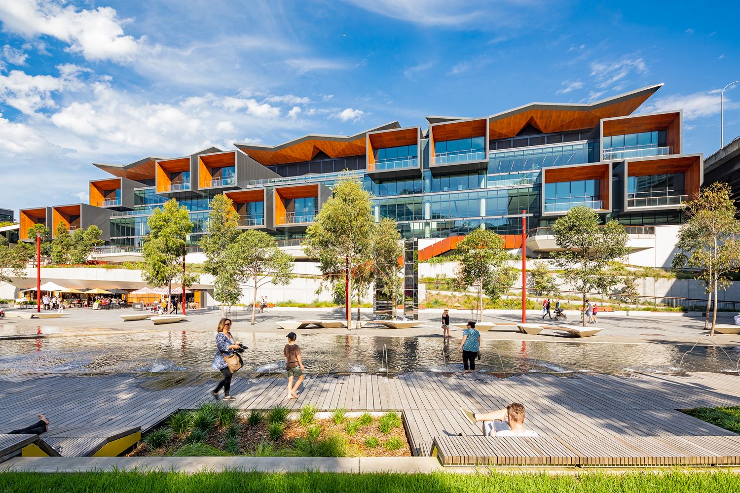 Visitors walk across a wet reflective plaza in front of a tiered convention building with orange wood-clad balconies and landscaped gardens.