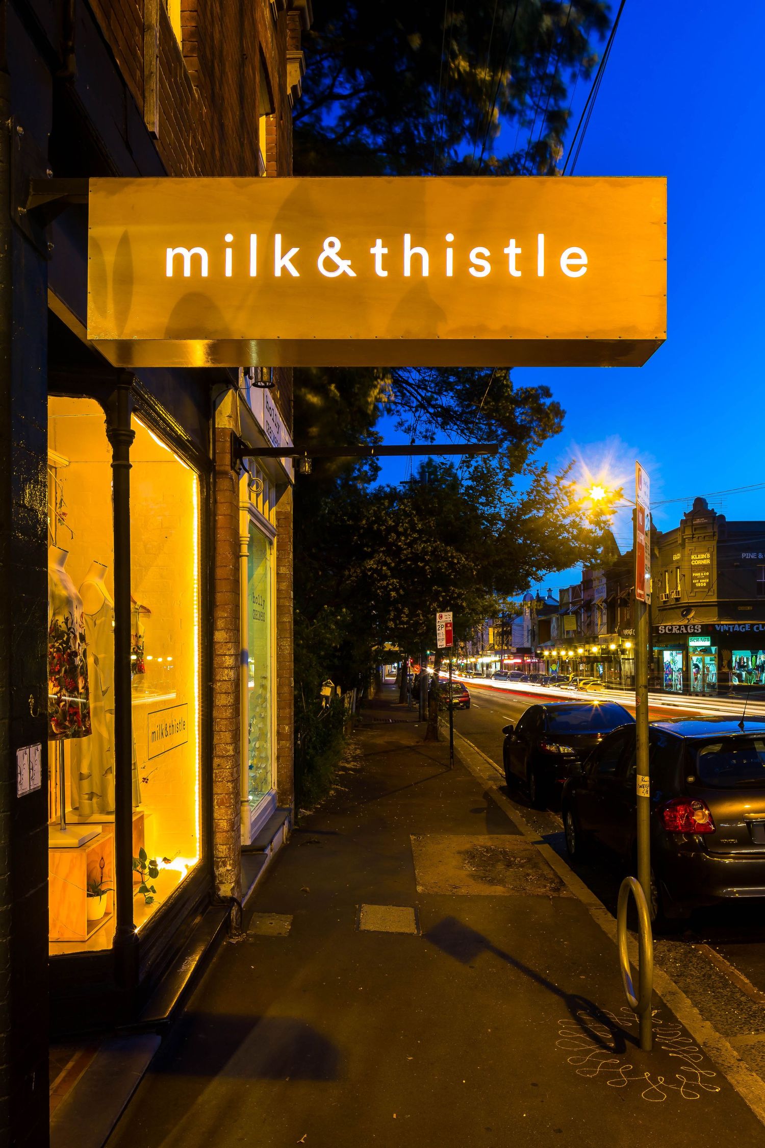 A glowing gold milk & thistle shop sign hangs over a lit storefront on a tree-lined street at dusk with passing car lights.