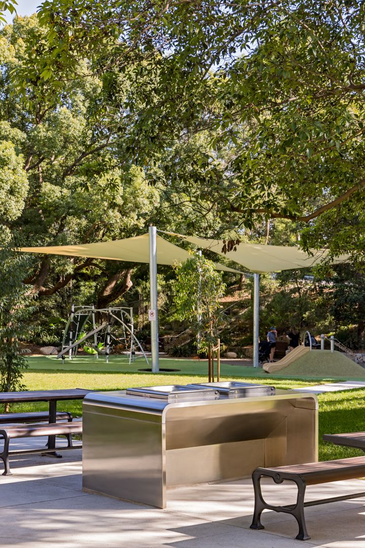 Stainless steel barbecue station and wooden benches beside a shaded park lawn with a playground and shade sail.