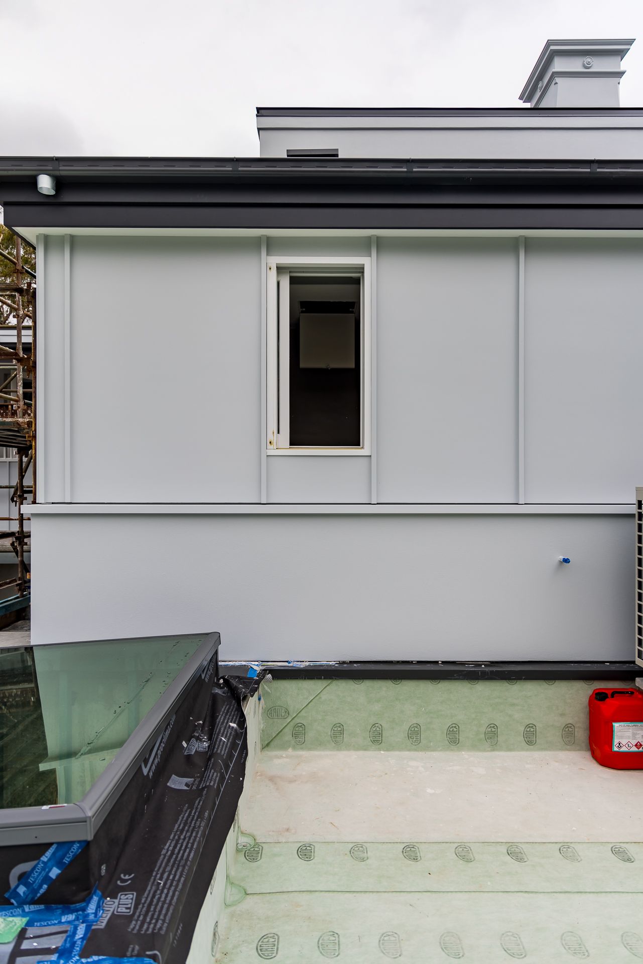 Side view of a grey clad building with a small window and chimney, with a skylight and green waterproof membrane on the roof terrace.