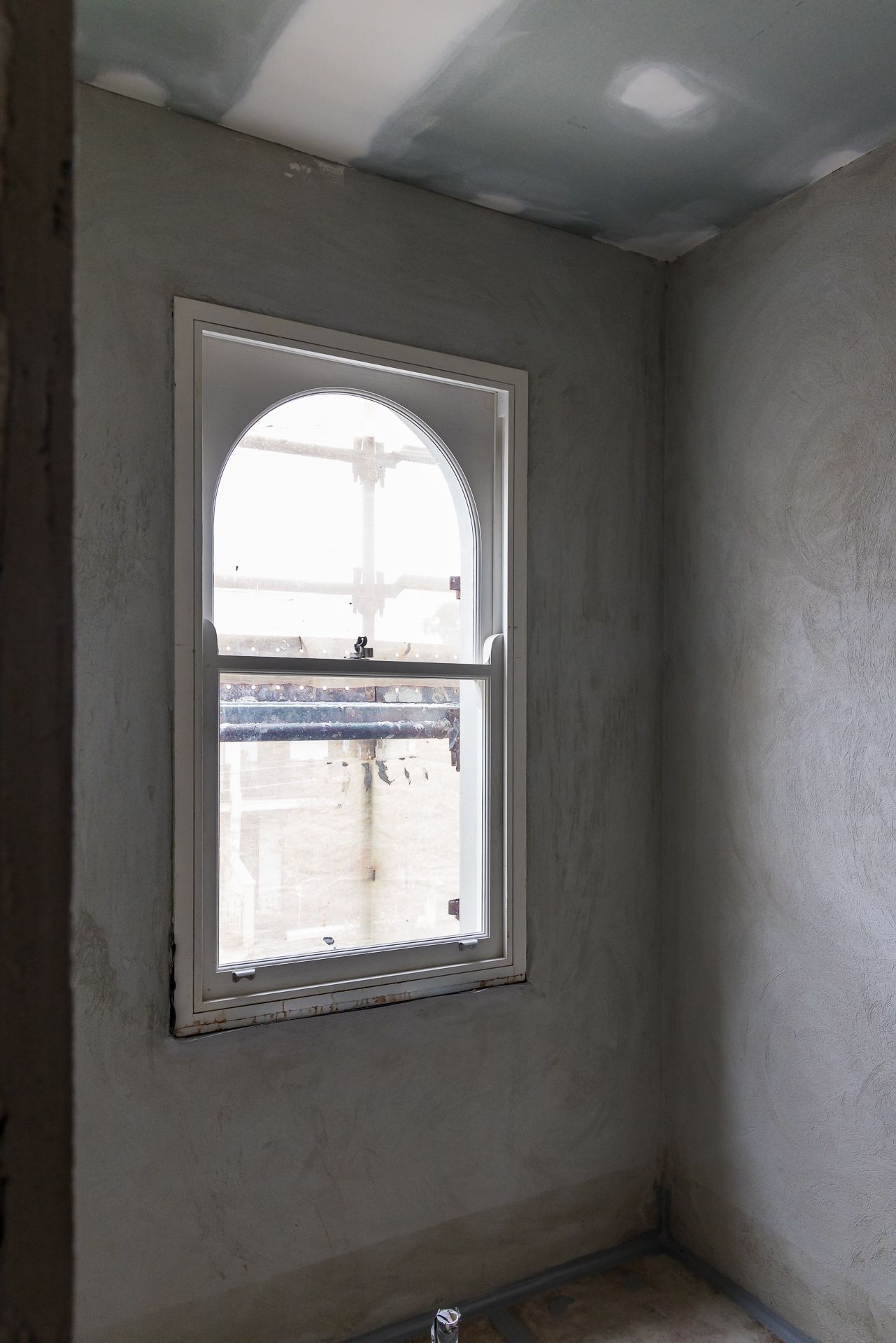 Arched-top timber sash window in a rough plastered room under renovation with unfinished ceiling plasterboard.