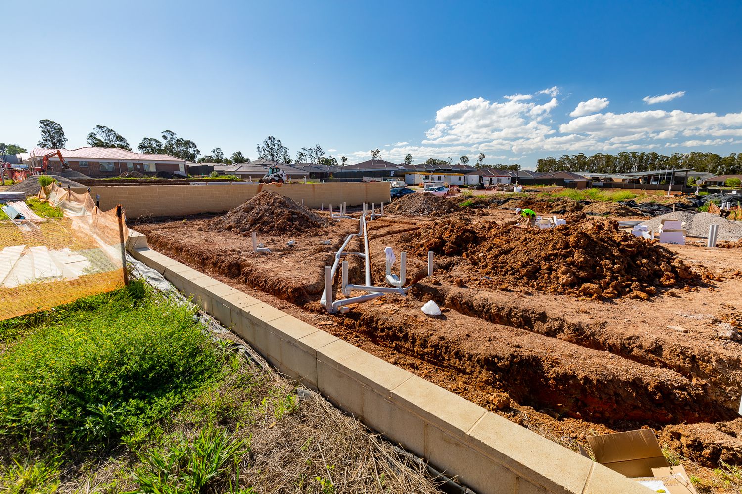 Excavated red soil and exposed plumbing pipes at a residential building site beside a concrete edge under a blue sky.