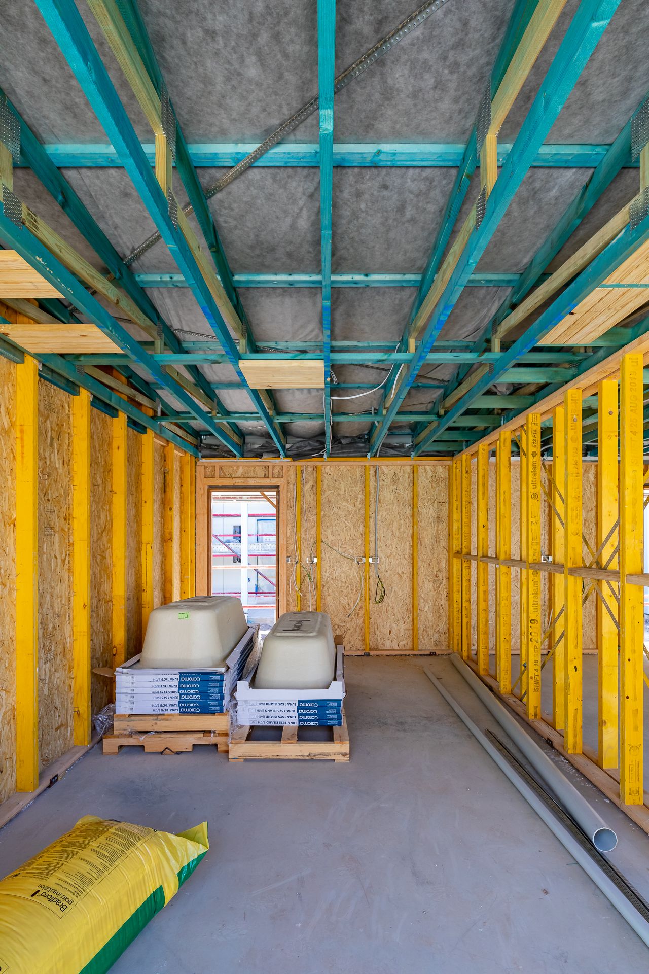 Timber-framed interior of a home under construction with teal steel ceiling joists, stacked bathtubs on pallets and a bag of insulation.