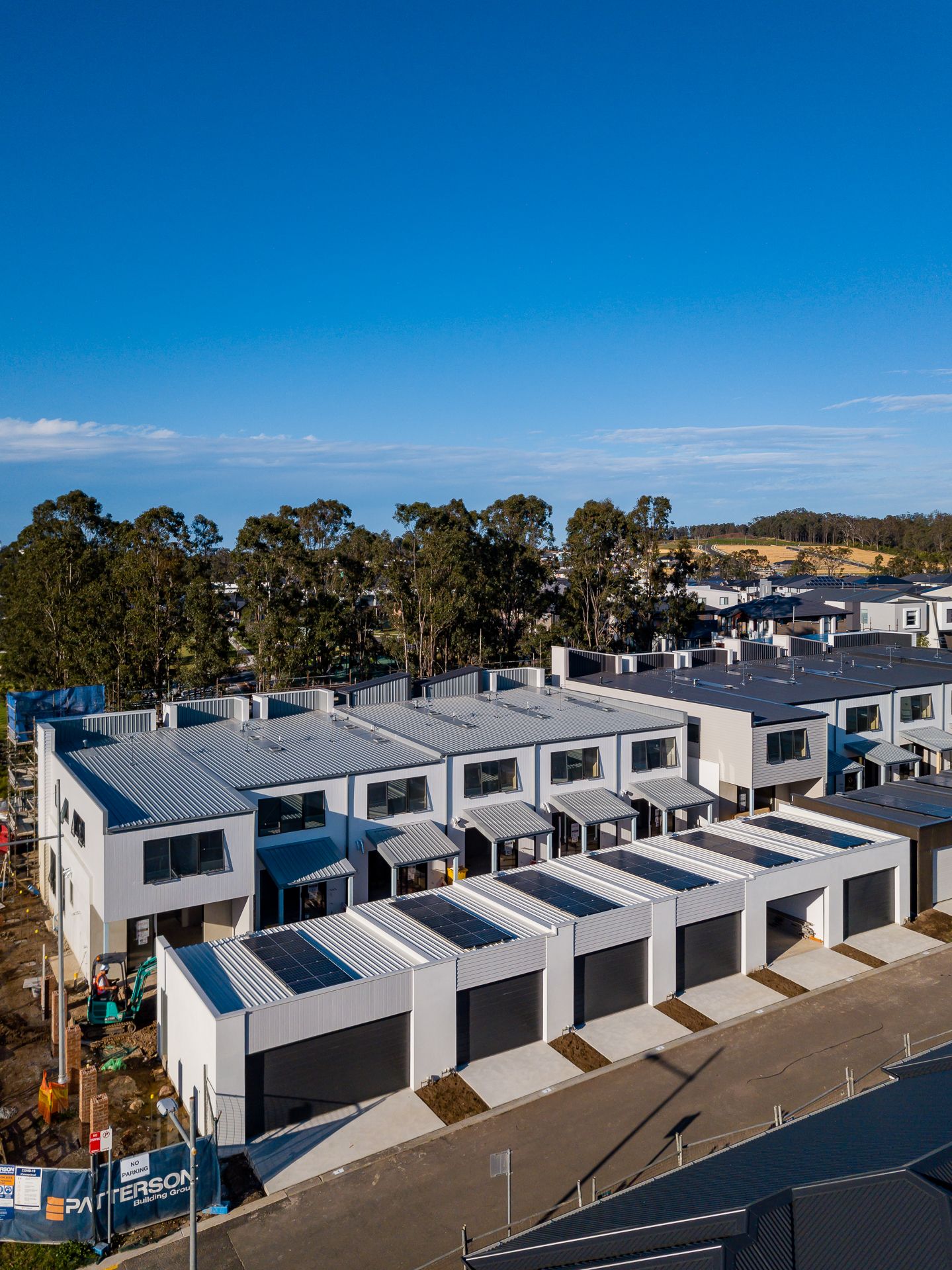 Drone view of newly built townhouses with solar panels, garages and tree-lined surroundings in a developing residential estate.