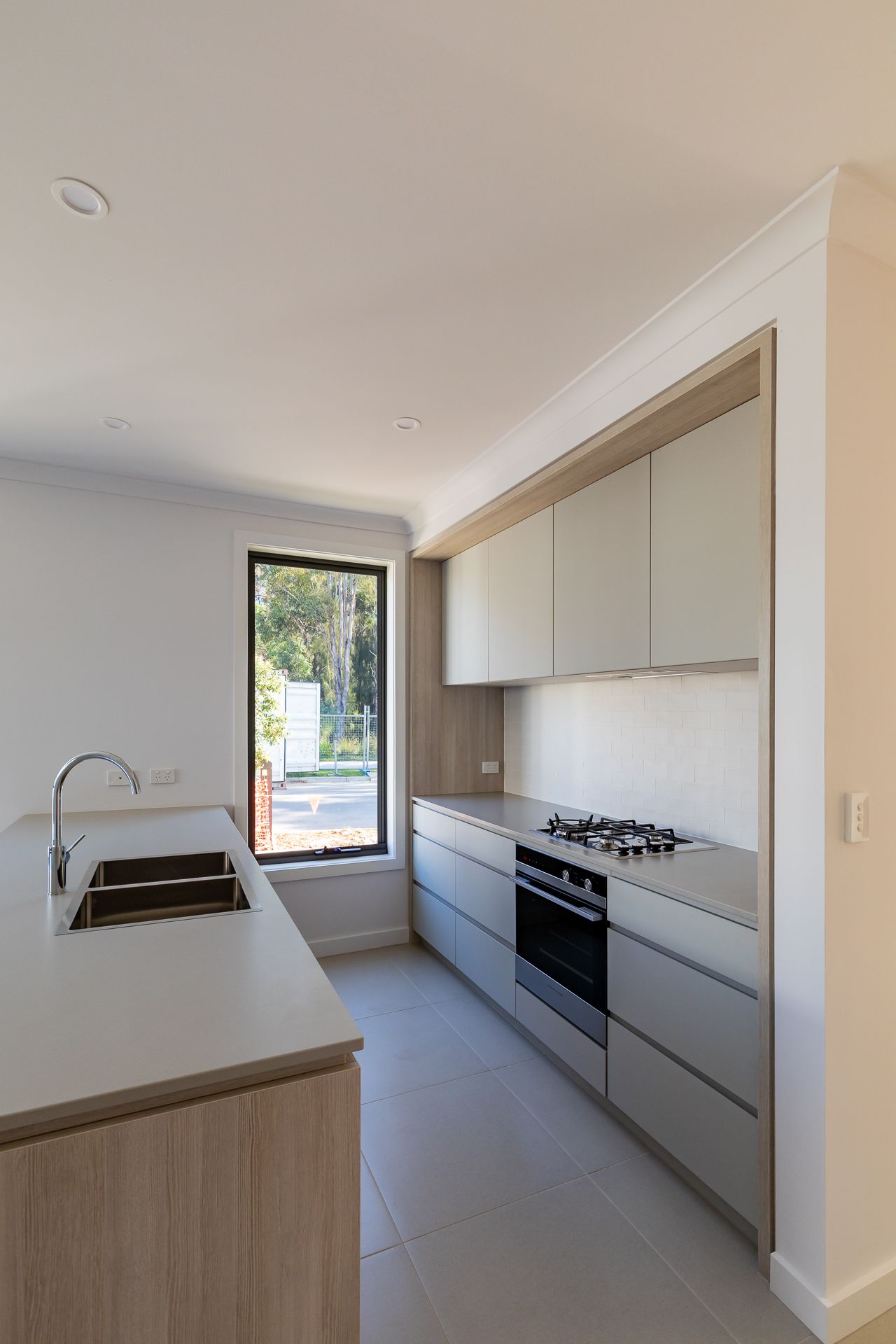 Modern kitchen with a double sink, timber-edged island, gas cooktop, wall oven and light cabinetry beside a tall window overlooking trees.