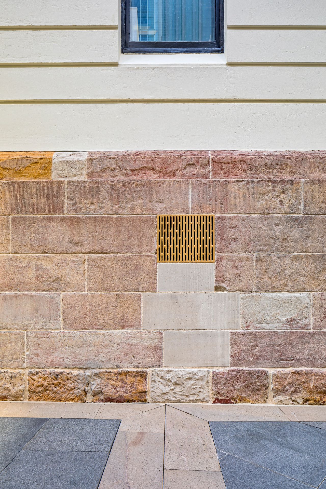 Sandstone block wall with a small yellow metal ventilation grate below a cream clapboard facade and window, above a paved stone pavement.
