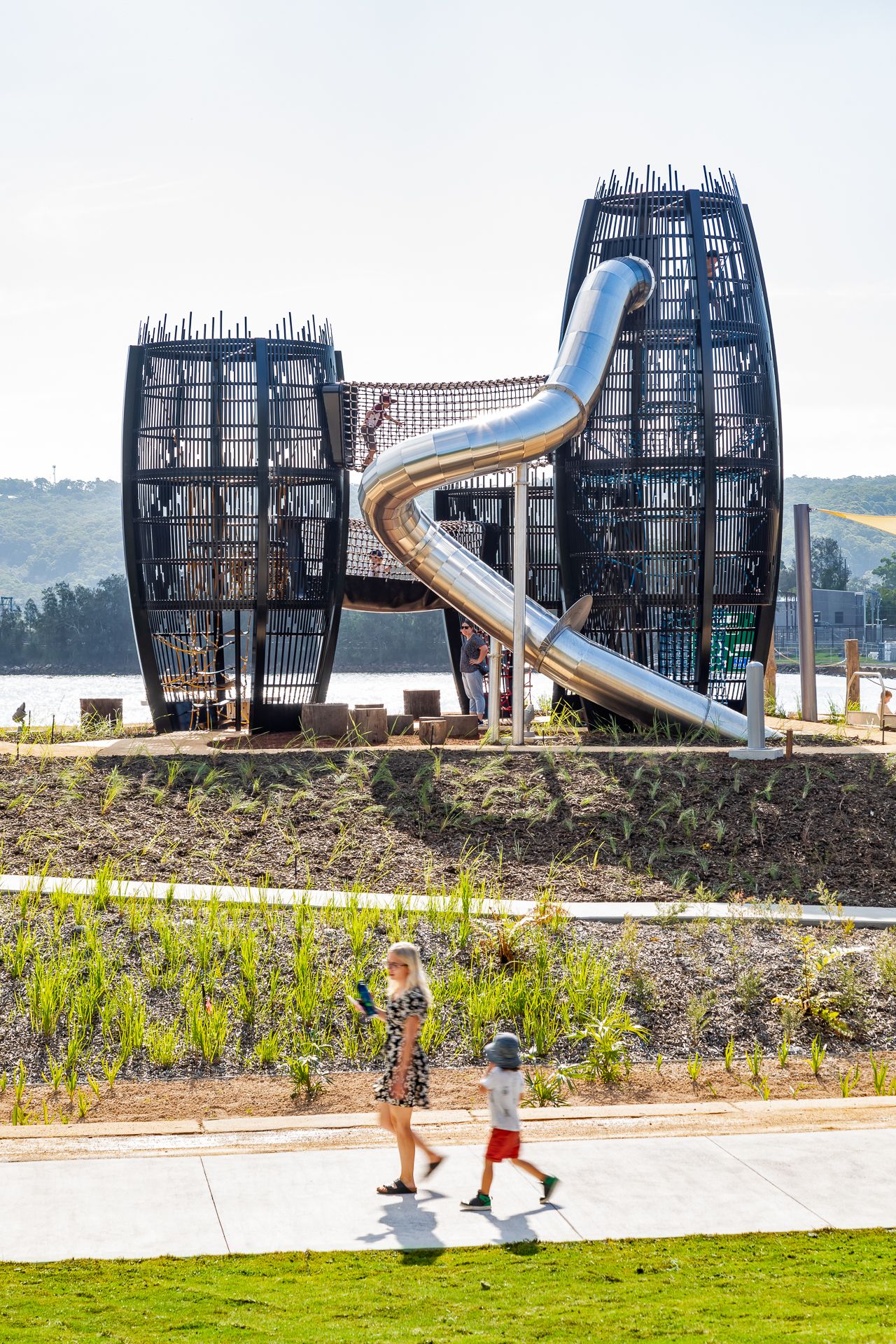 A woman and young child walk along a path in front of towering black play structures with a curved metal slide beside the water.