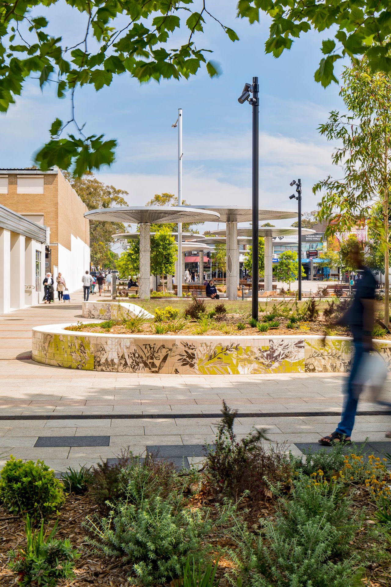 Native plantings and a curved mosaic garden wall frame a modern public plaza with tall light poles and distant umbrella-style canopies.