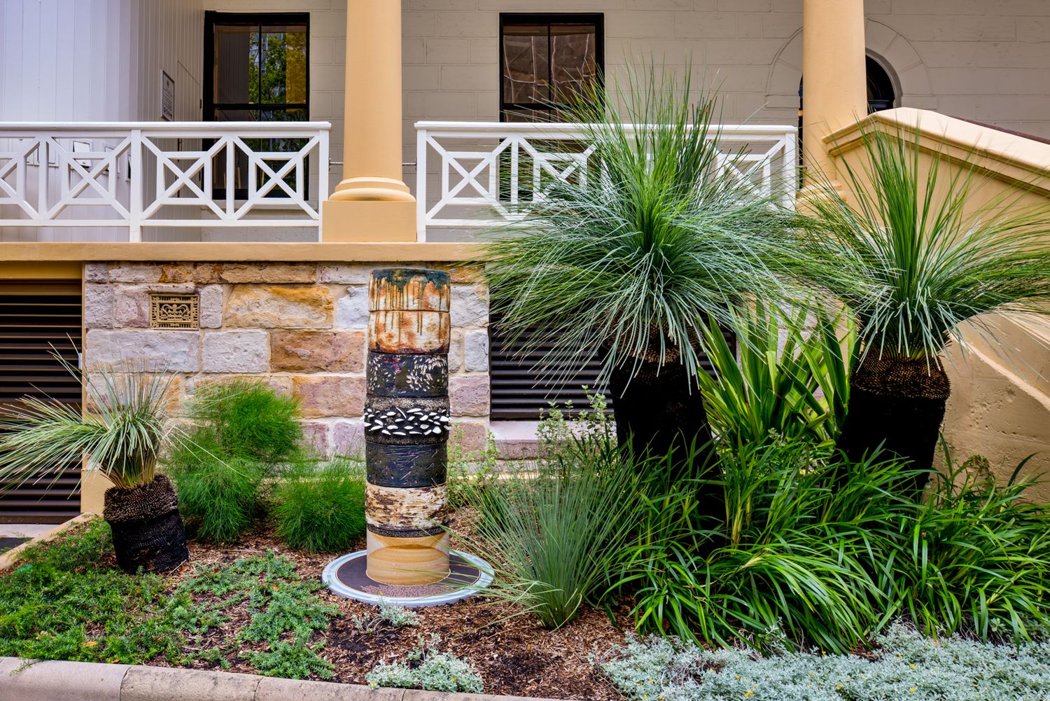 Sculptural totem column with layered stone and ceramic textures stands in a landscaped garden of grasses beside a sandstone building.