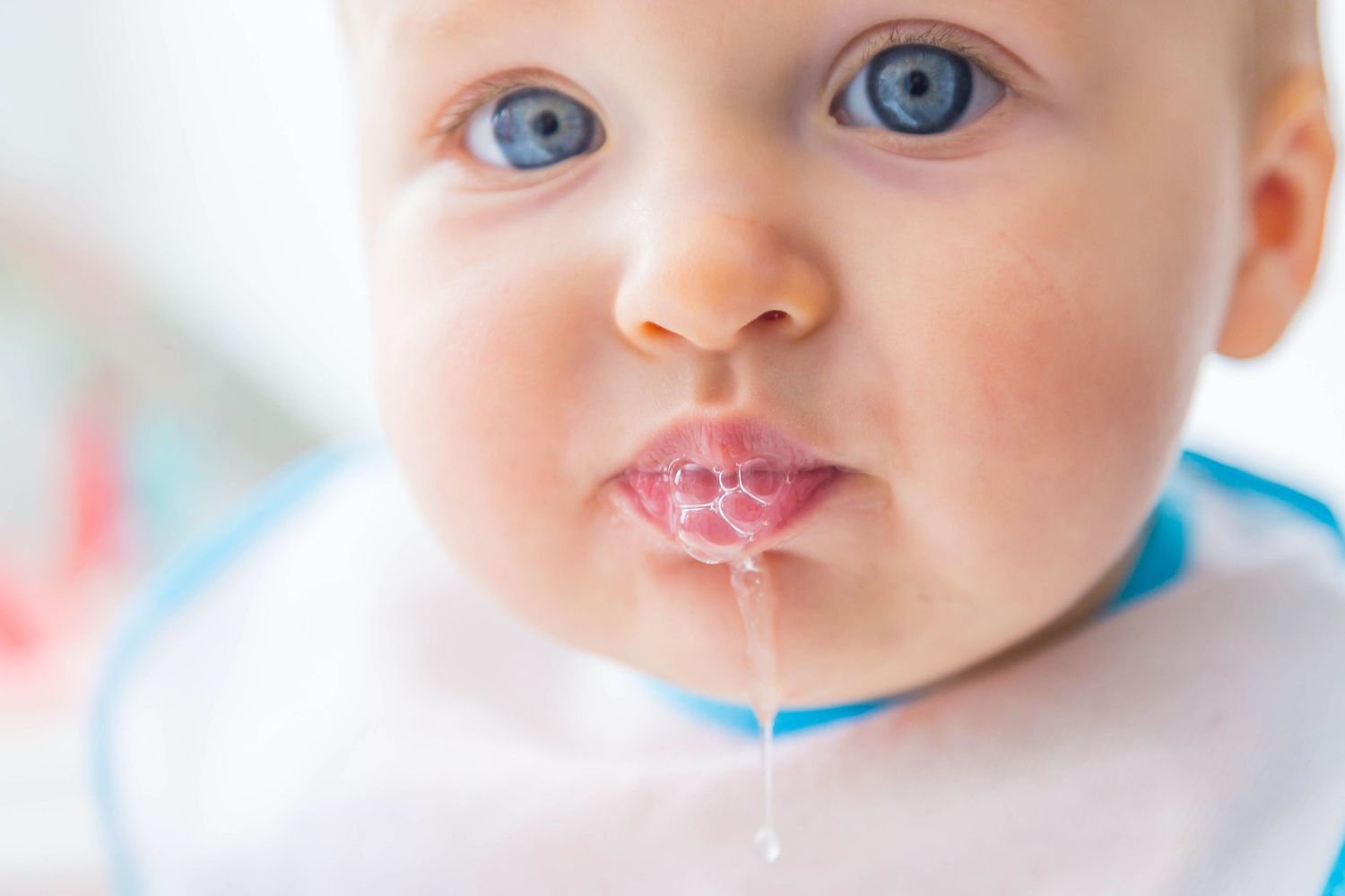 Close-up portrait of a blue-eyed baby in a bib with drool dripping from its lips.