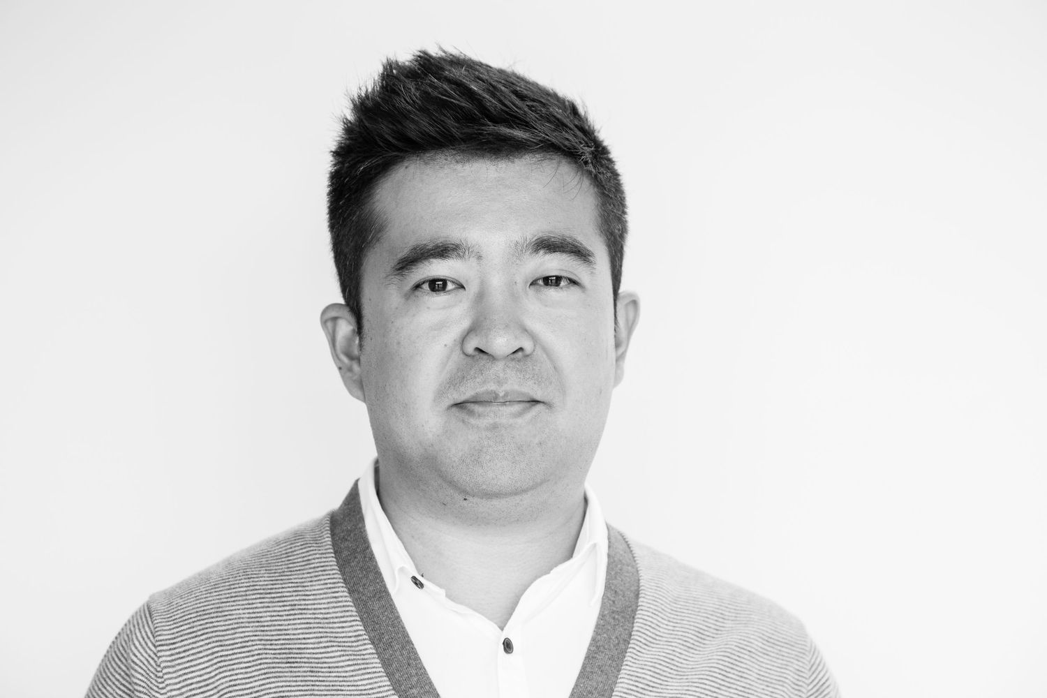 Black and white portrait of a man with short spiky hair wearing a cardigan over a collared shirt against a plain white background.
