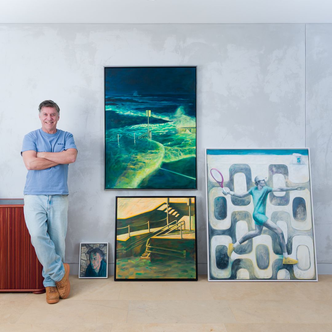 Man in a blue t-shirt and jeans stands with arms crossed beside colorful paintings of ocean waves, a staircase and a tennis player.