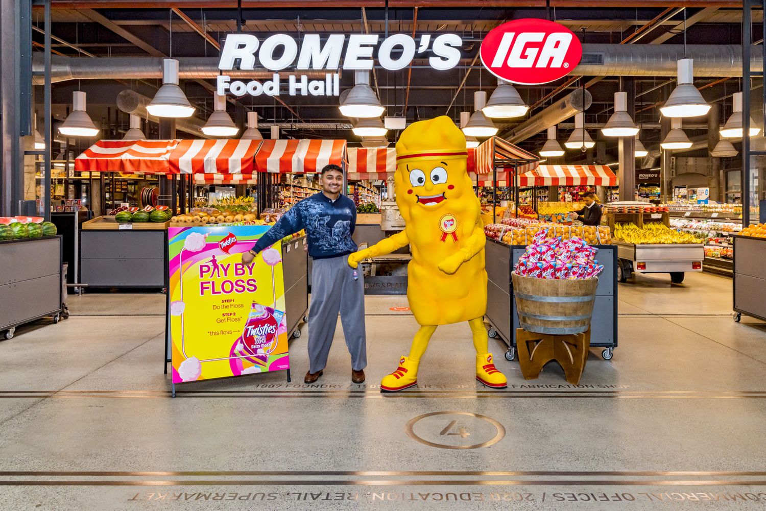 A man stands beside a yellow dental floss mascot and a Pay It Floss sign inside Romeo's IGA Food Hall.