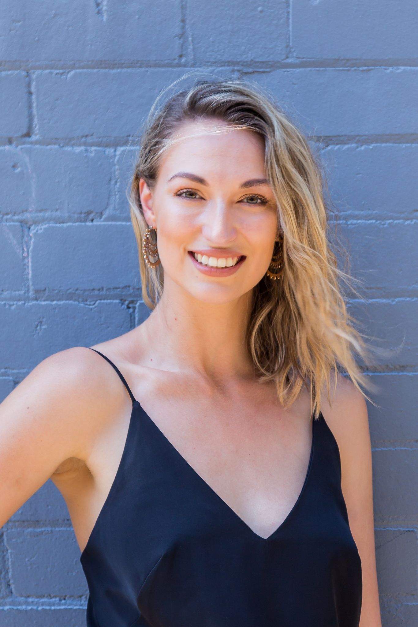 A smiling woman with wavy blonde hair and gold earrings wears a black camisole while posing against a painted gray brick wall.