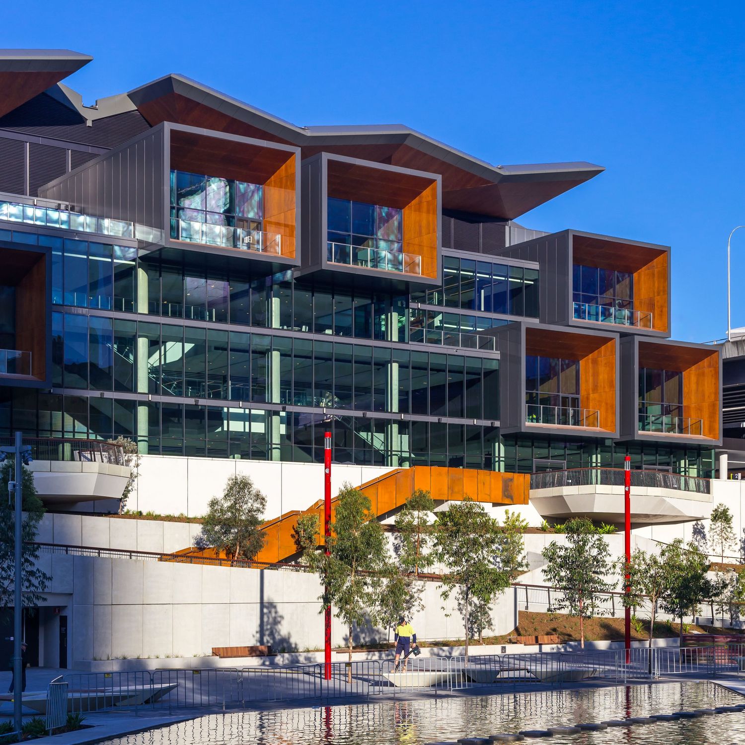 Modern waterfront building with glass facade, timber-clad balconies and an orange staircase beside landscaped trees and a reflective pool.