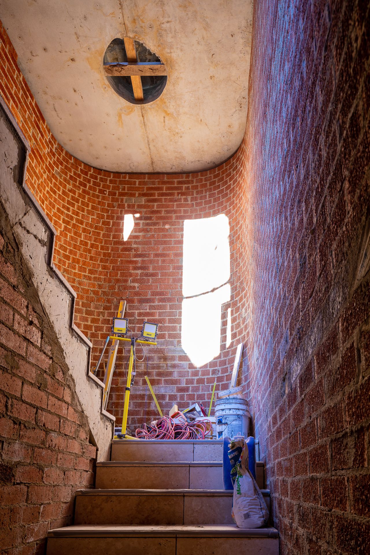 Brick-walled stairwell under construction with a curved concrete ceiling, bright window opening, survey equipment and cables on the steps.
