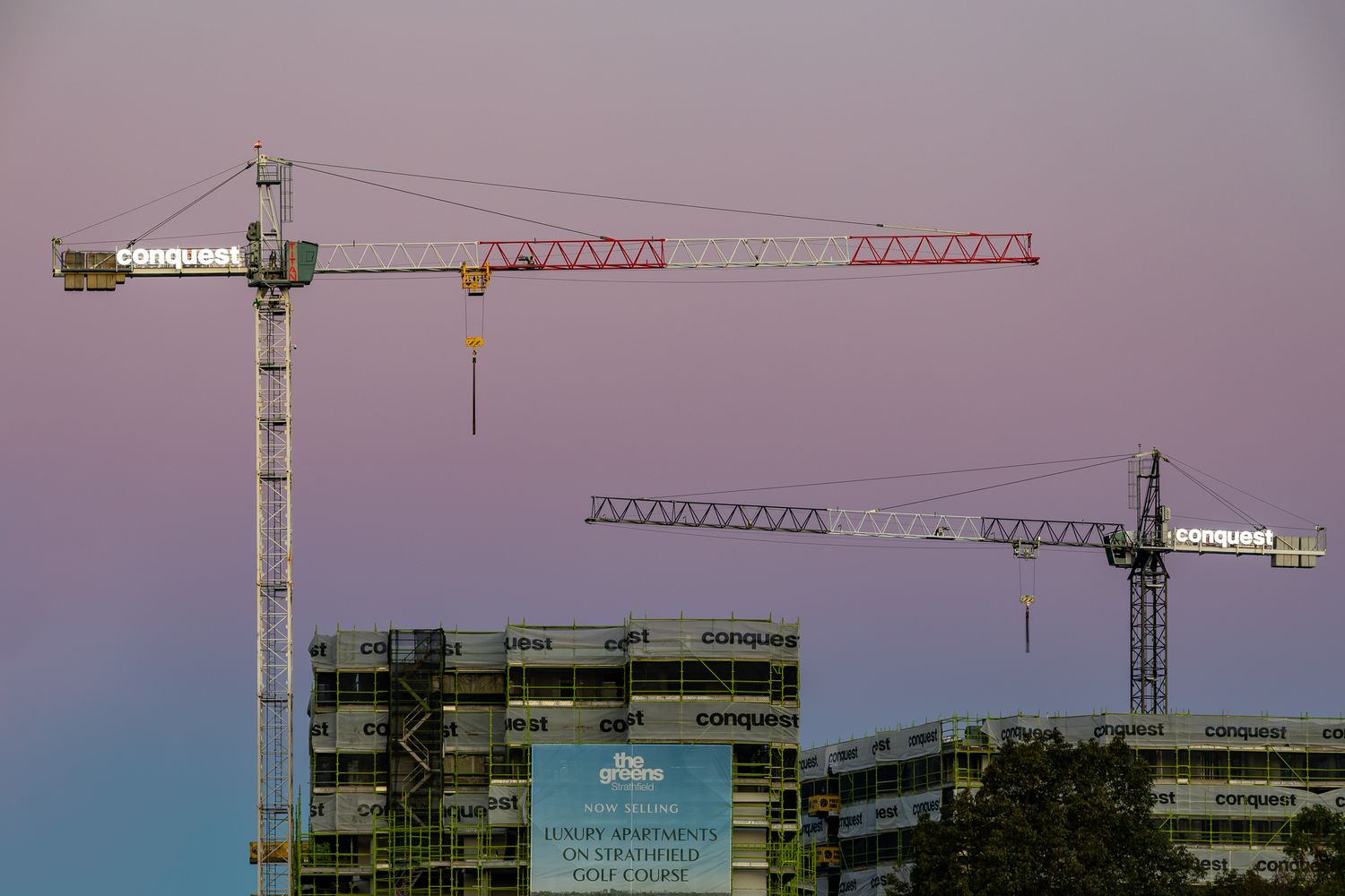 Construction cranes and an unfinished apartment building silhouetted against a pink and purple dusk sky.