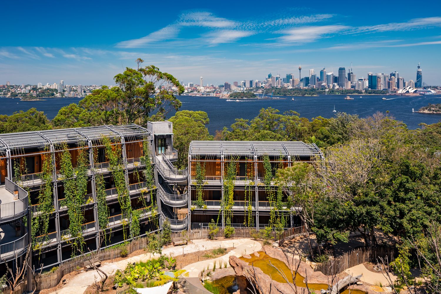 Aerial view of a modern multi-level building draped in climbing greenery with the Sydney skyline and harbour in the background.