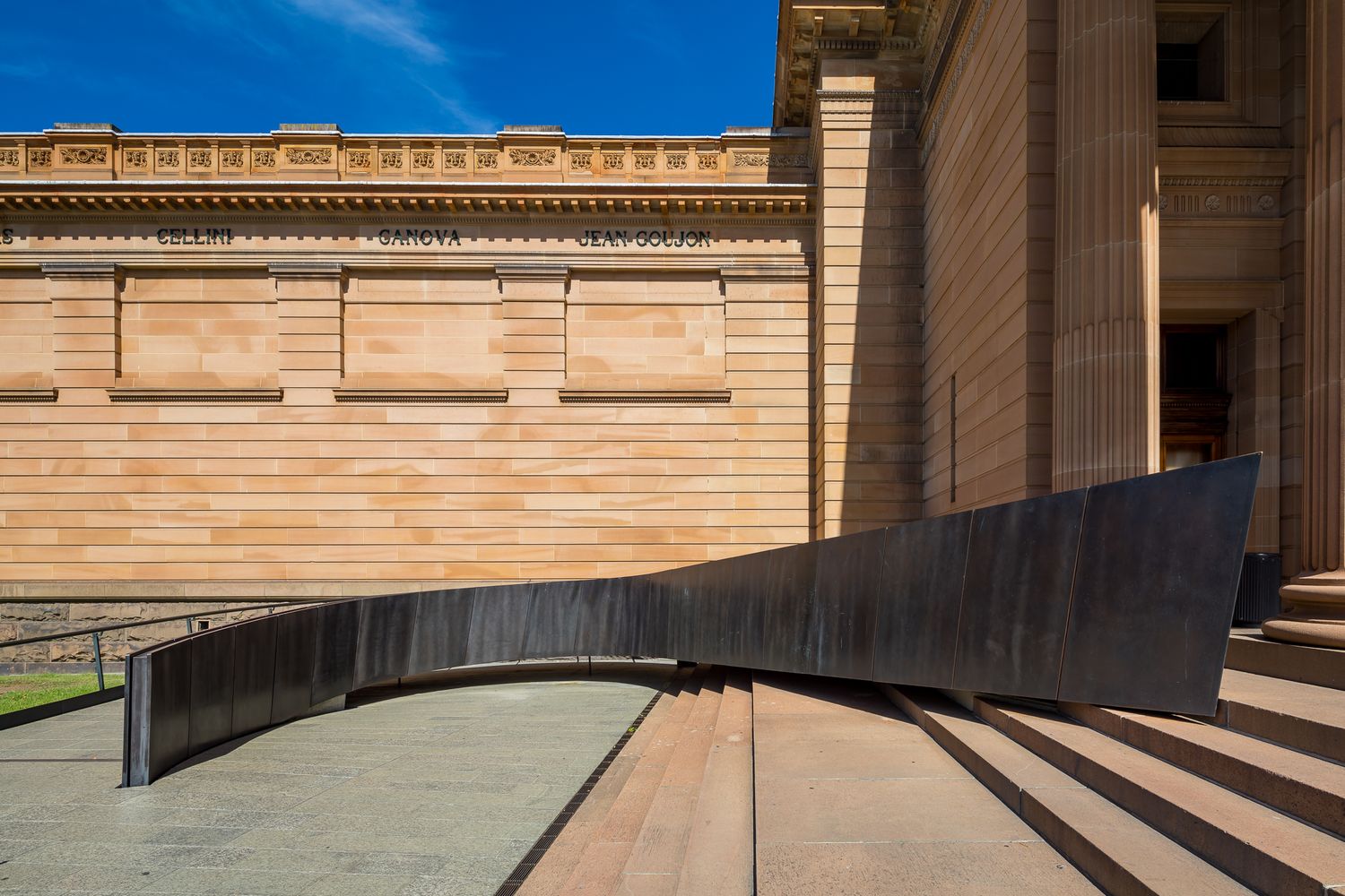 Curved dark metal sculptural bench in front of a sandstone heritage building with columns and steps under a blue sky.