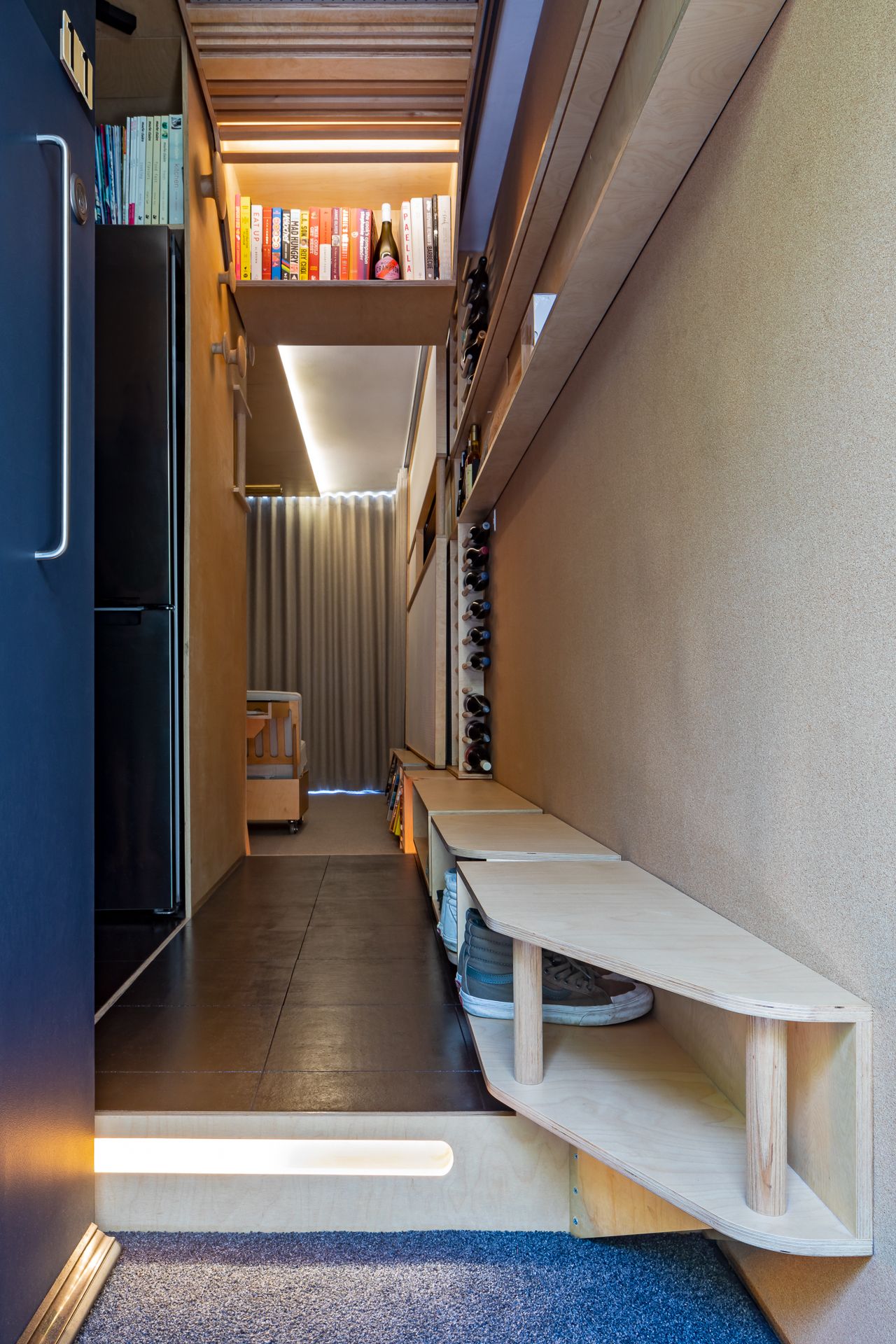 Narrow apartment entry hallway with plywood shoe bench, built-in wine racks, raised dark floor and overhead book shelf.