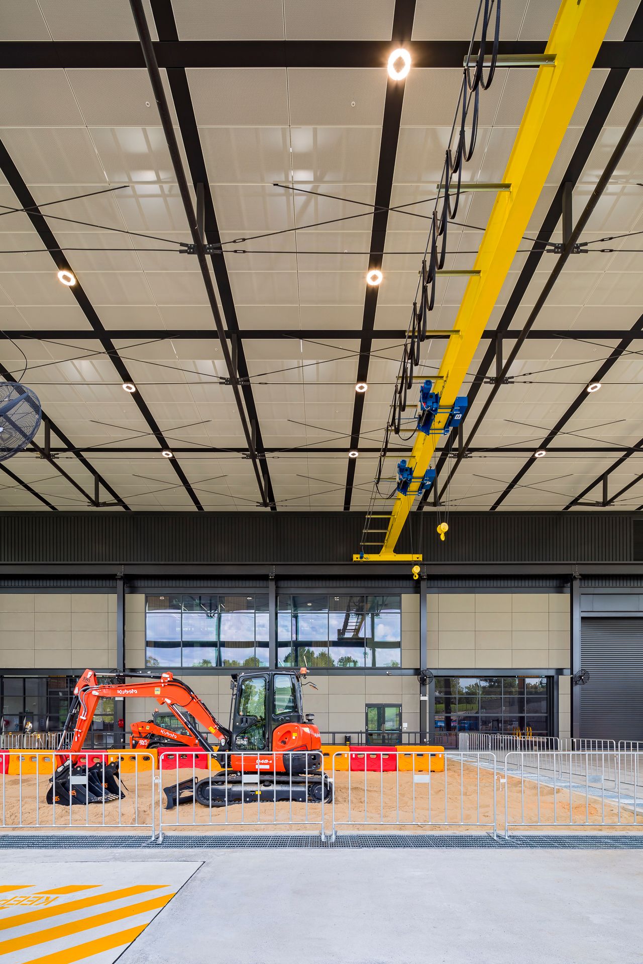Orange mini excavator parked behind safety barriers inside a large training workshop with a yellow overhead gantry crane and high ceiling.