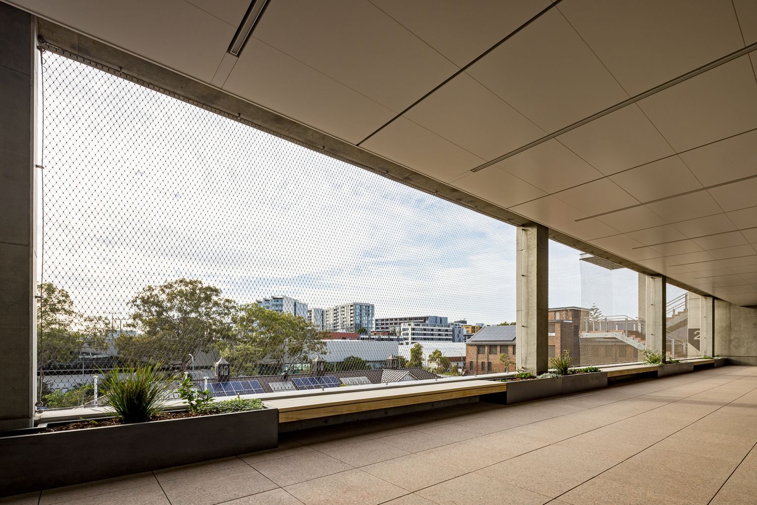Wide covered outdoor terrace with tiled flooring, planters and a bench looking out over trees and city buildings through protective mesh.
