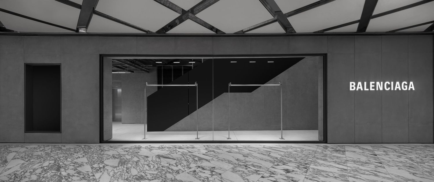 Minimalist Balenciaga boutique entrance with large glass windows, clothing racks, a geometric ceiling and polished marble floors.