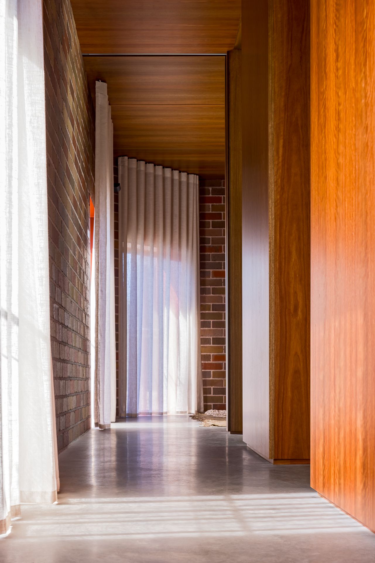 Modern hallway with polished concrete floors, exposed brick walls, warm timber paneling and sheer curtains glowing with natural light.
