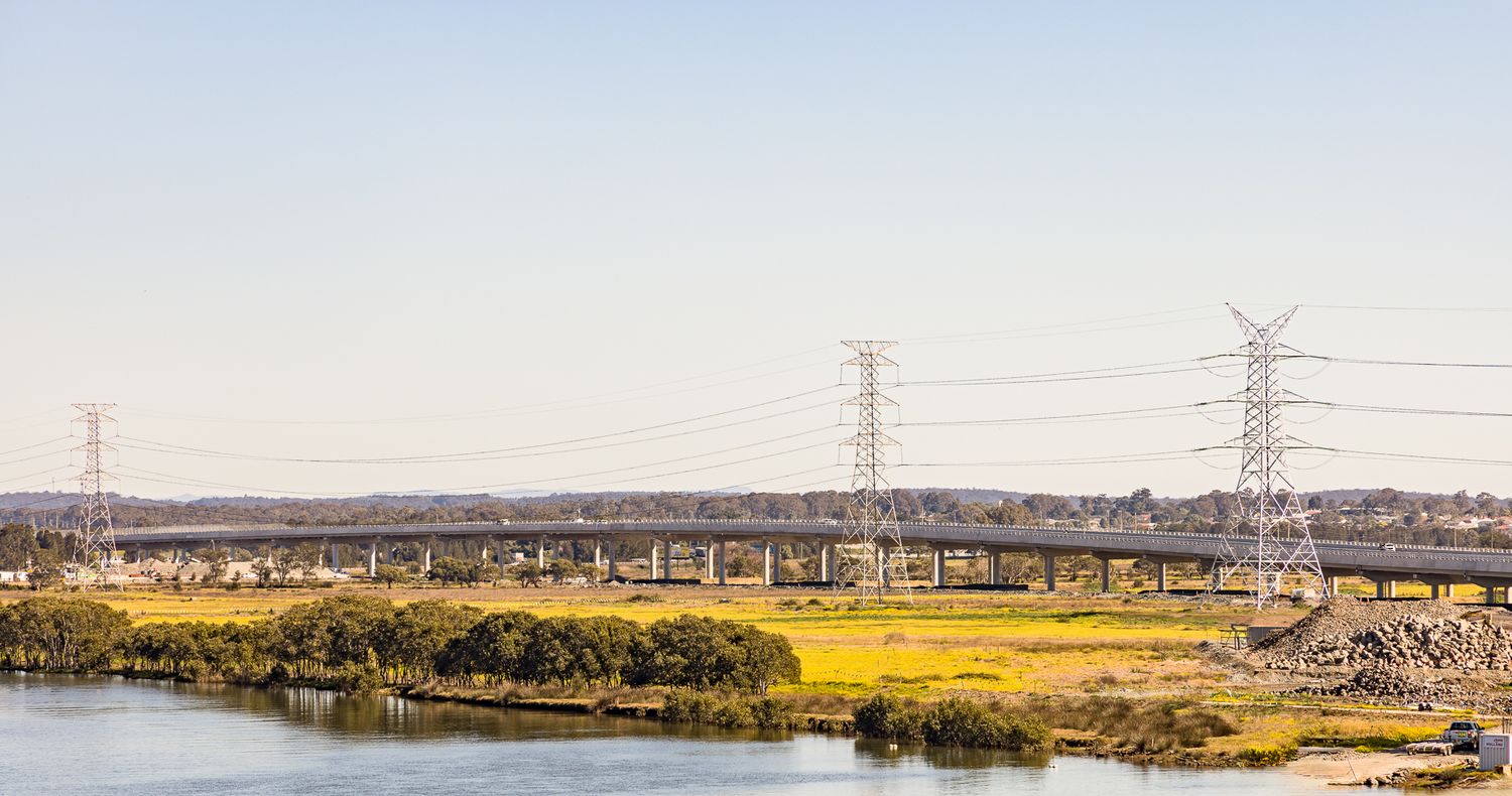 Long elevated motorway bridge spanning wetlands and a waterway with electricity pylons beneath a hazy sky.