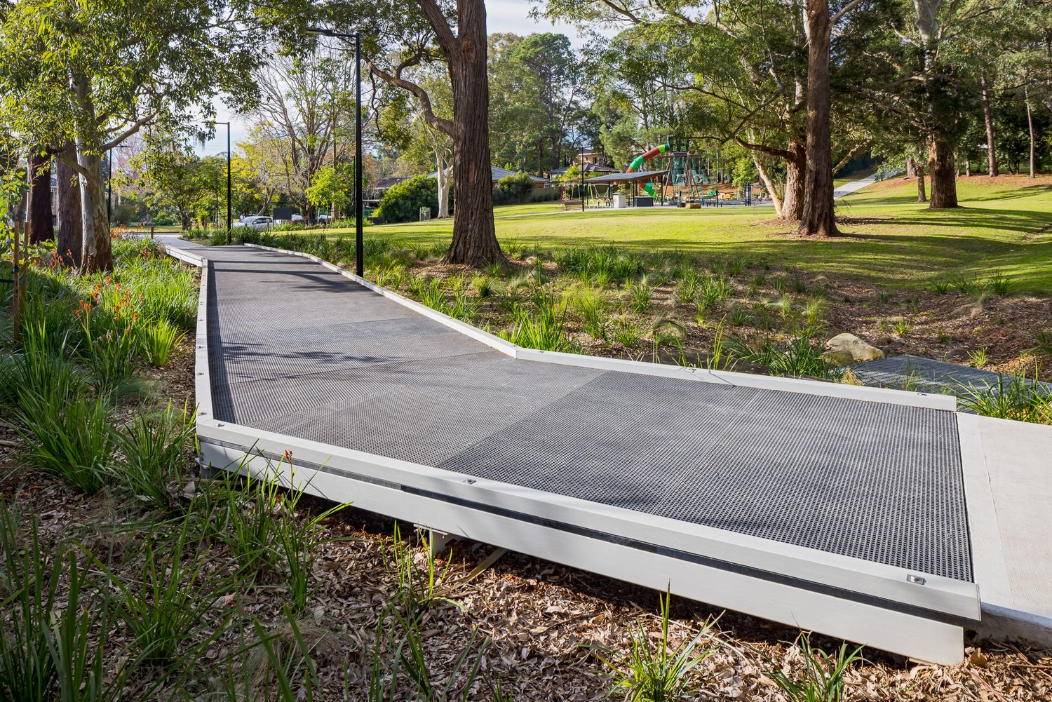 Modern ramped concrete and metal pathway with grated surface cuts through native garden beds beside tall gum trees in a park.