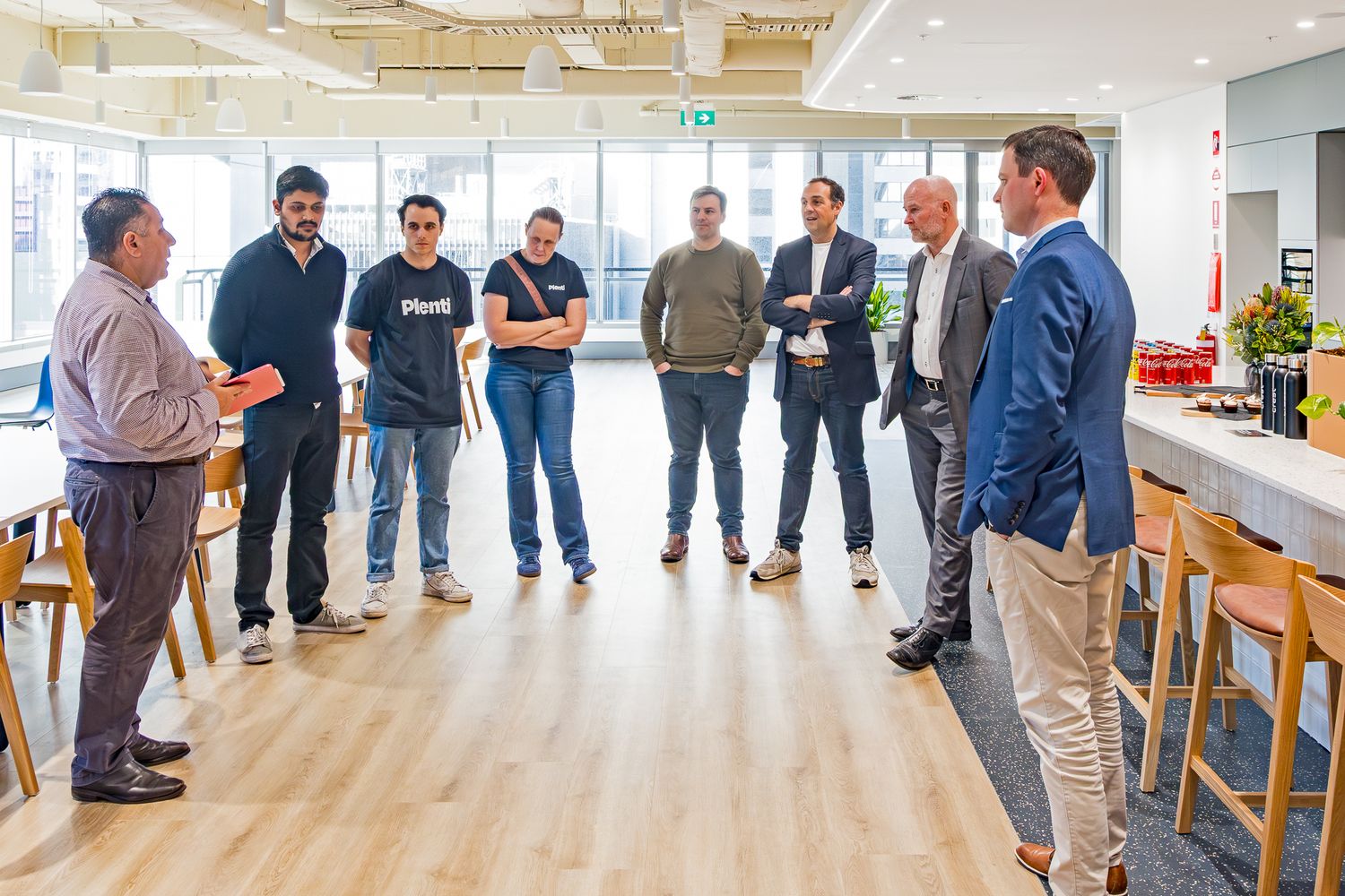 Group of coworkers hold an informal standing meeting in a bright office break area with wooden stools and a kitchen counter.