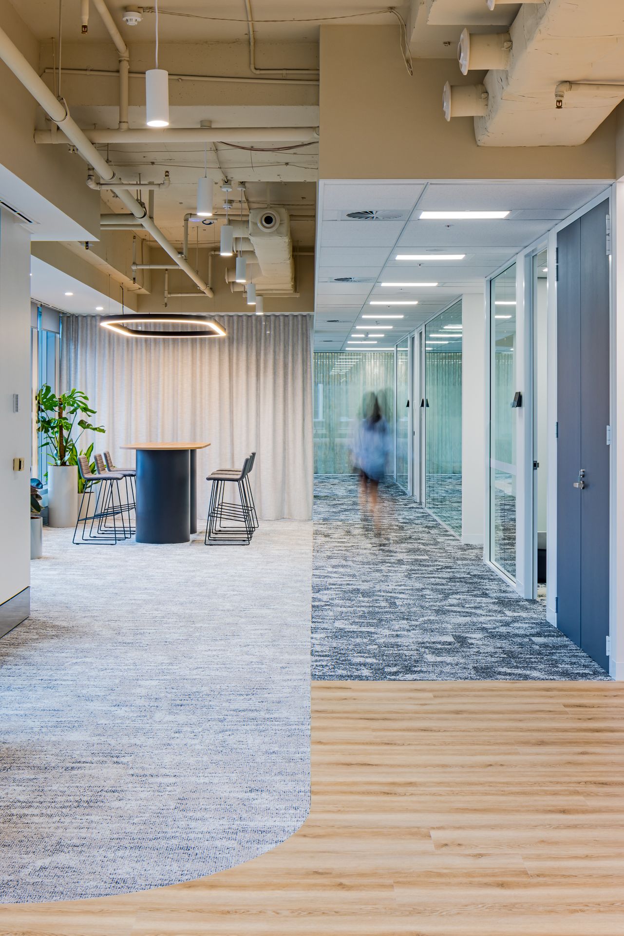 Open office corridor with mixed carpet and timber flooring, a high table with stools, glass-walled meeting rooms and a person walking past.