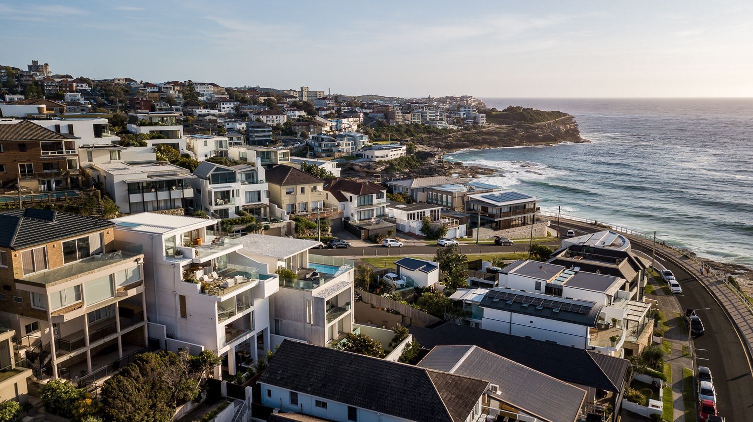 Coastal neighbourhood of modern homes with balconies overlooking the ocean and a winding seaside road.
