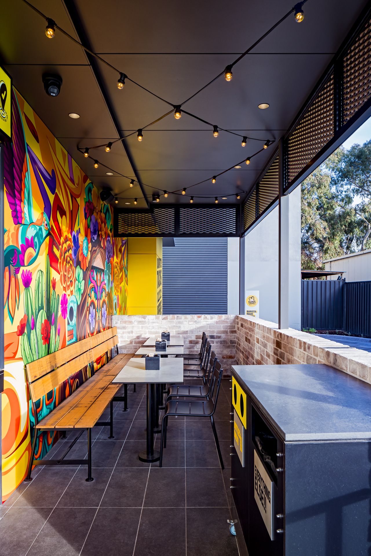 Covered outdoor dining area with timber benches, string lights, brick walls and a bright floral mural at a Mexican restaurant.