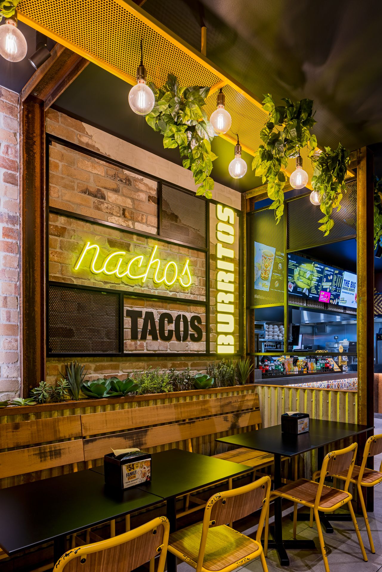 Dining booth with neon nachos sign, tacos and burritos wall graphics, exposed brick, pendant lights and trailing green plants.