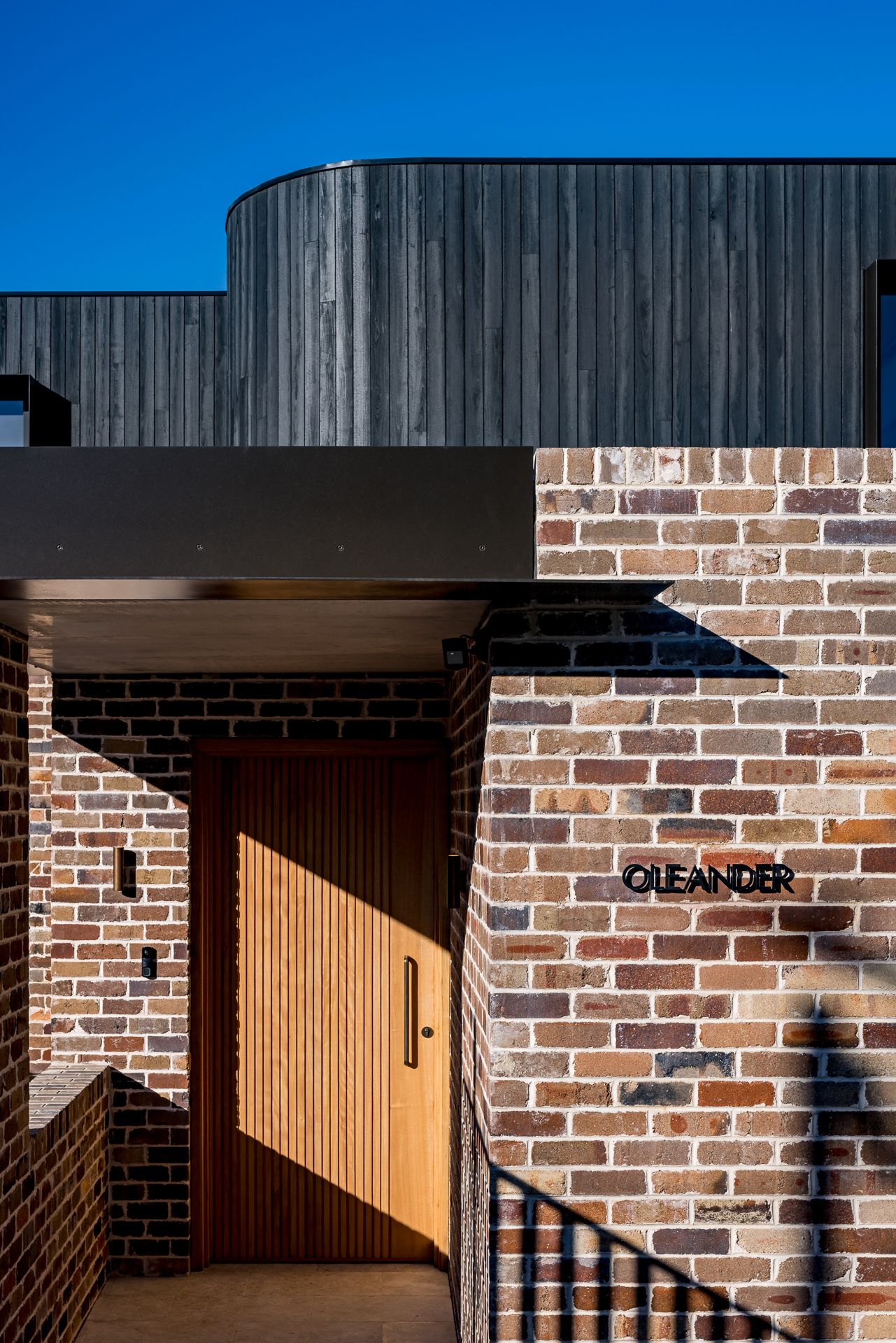 Entrance detail of a brick home with a timber front door, black steel canopy, curved charred timber cladding and an Oleander sign.
