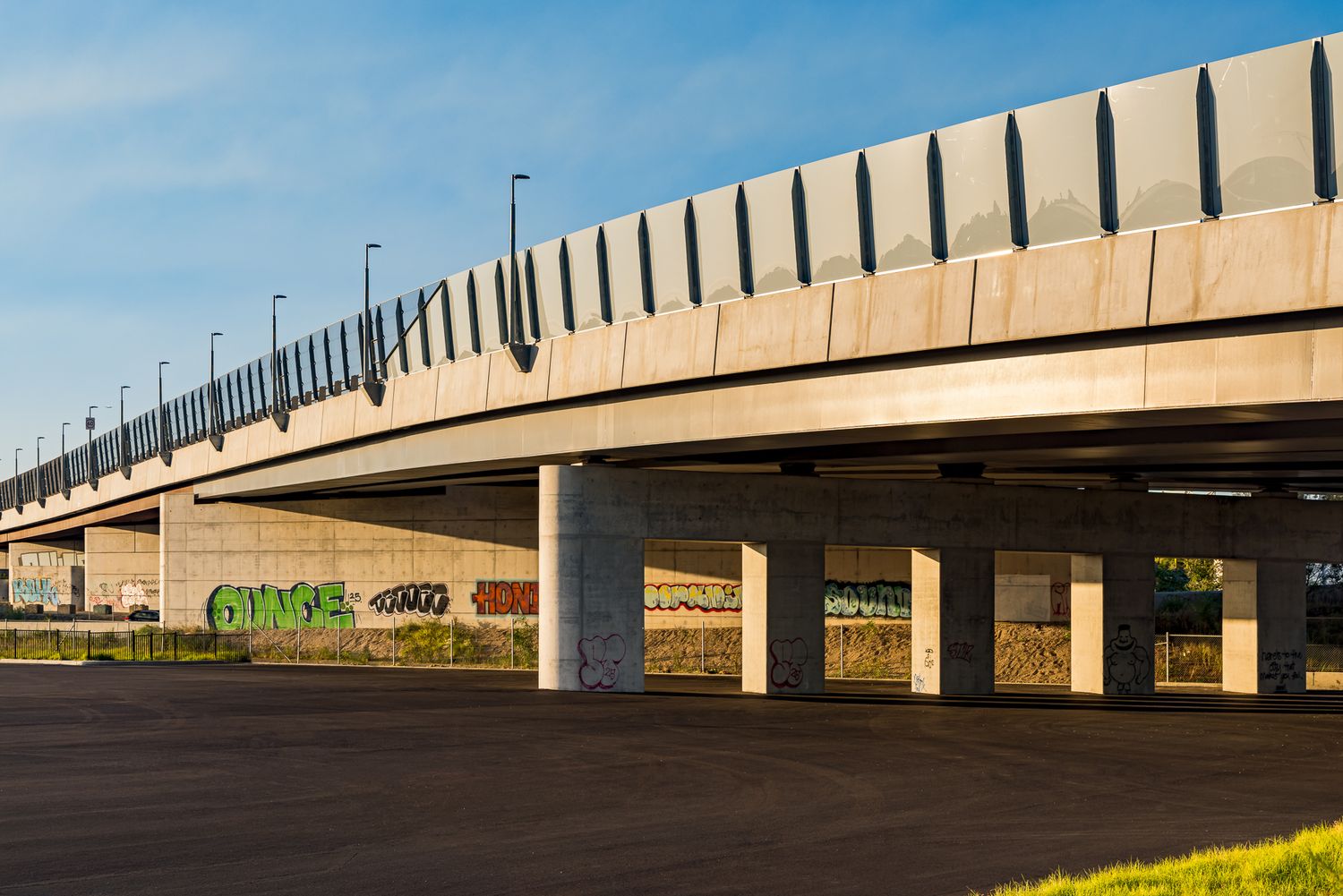 Concrete elevated roadway bridge with colorful graffiti on the support pillars and an empty paved area below under a blue sky.