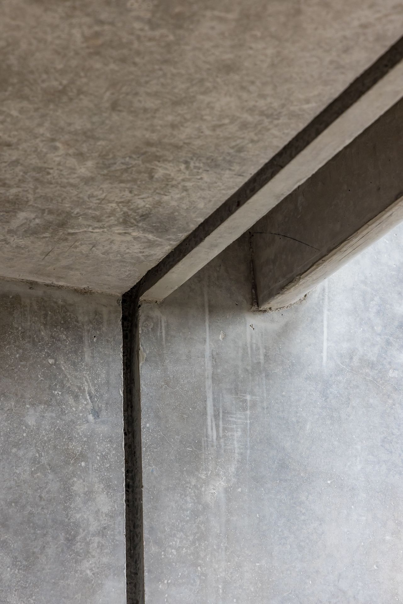 Close-up of a concrete ceiling and wall corner with a thin dark expansion joint and subtle texture and water stains.