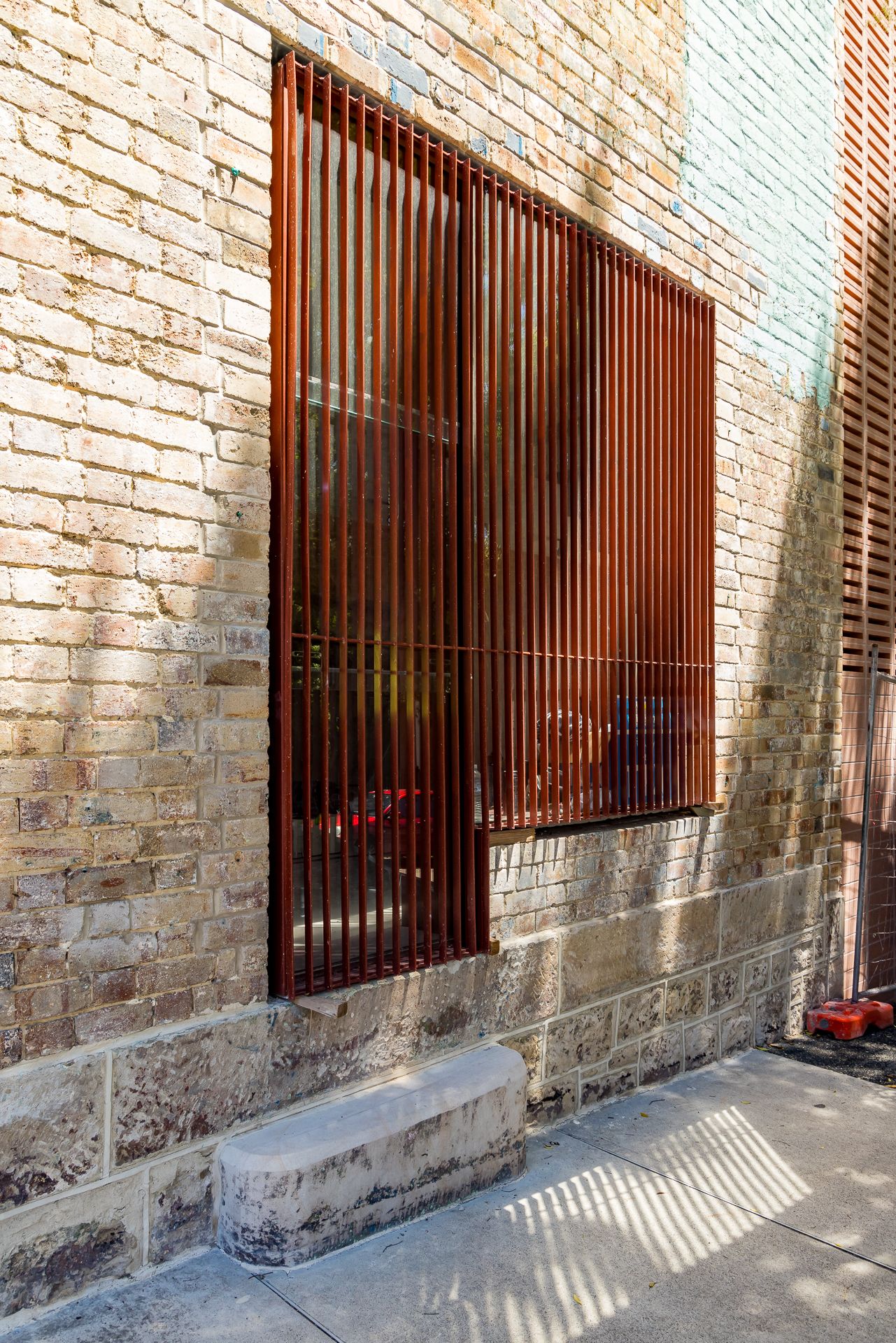 Rust-colored vertical steel slat screen over a window in a sandstone and brick wall with a stone step below.