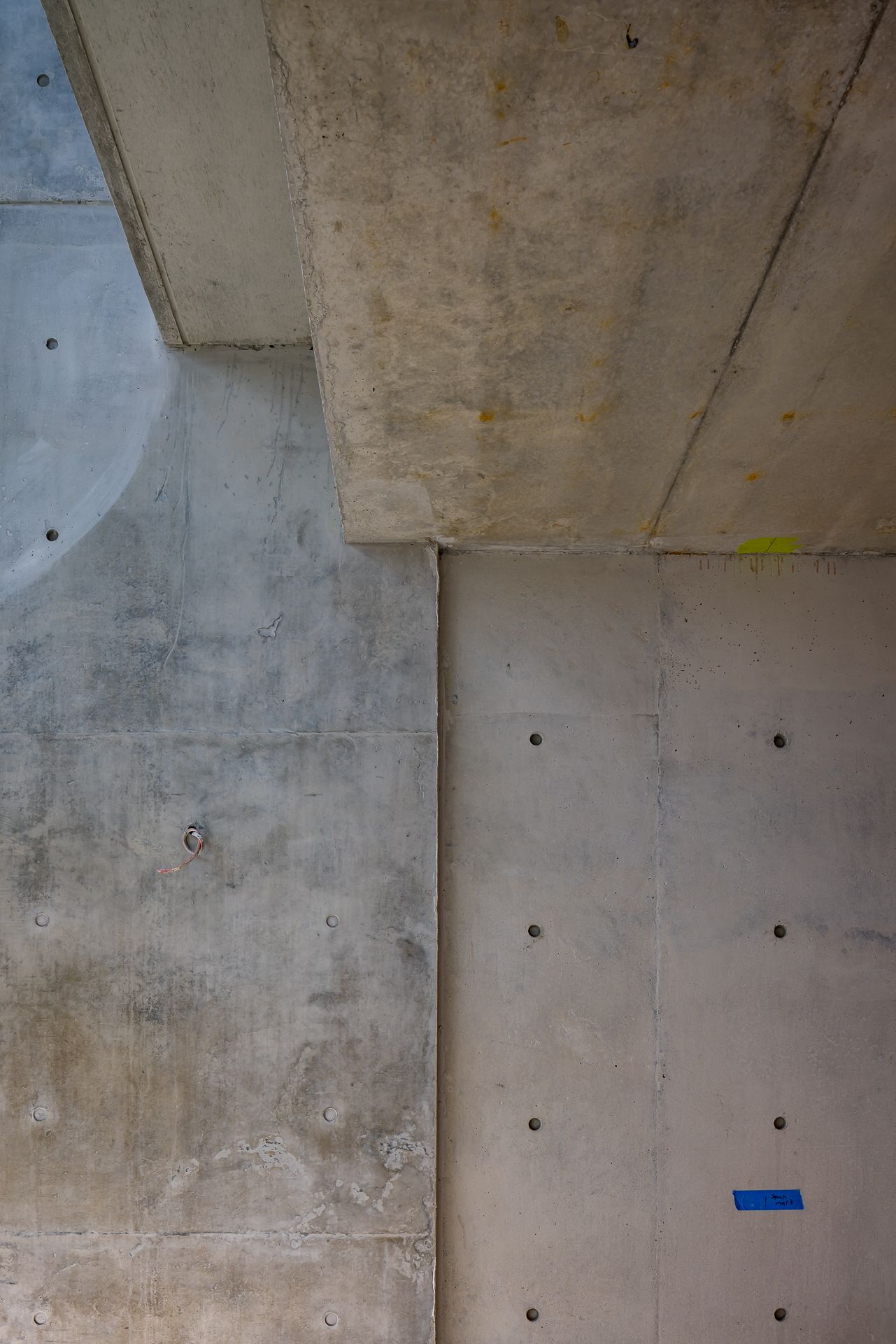 Board-formed concrete wall corner with evenly spaced tie holes, weathered stains and a small blue sticker mark.