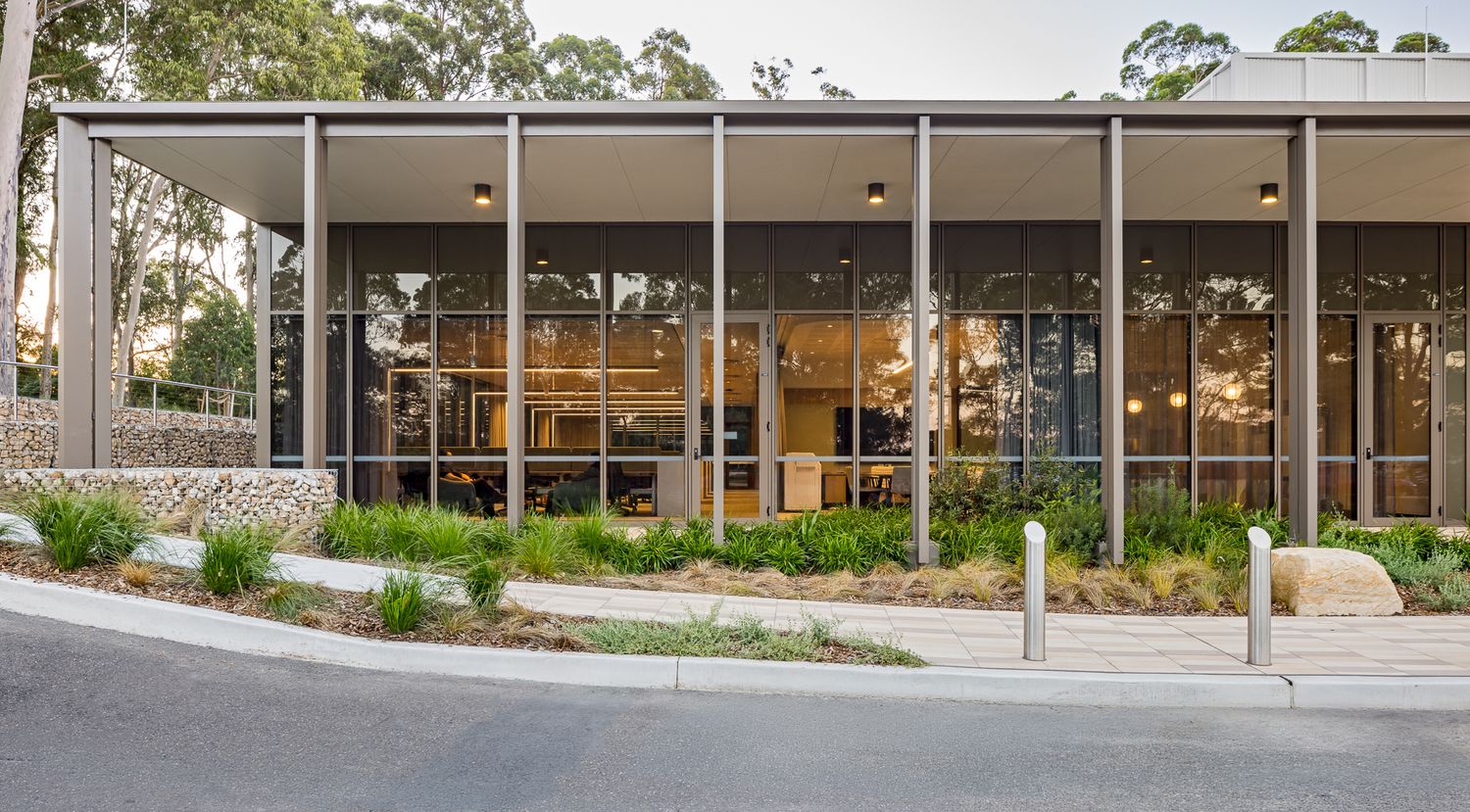 Single-storey modern building with floor-to-ceiling glazing, slender steel columns and a flat canopy roof beside native garden beds.