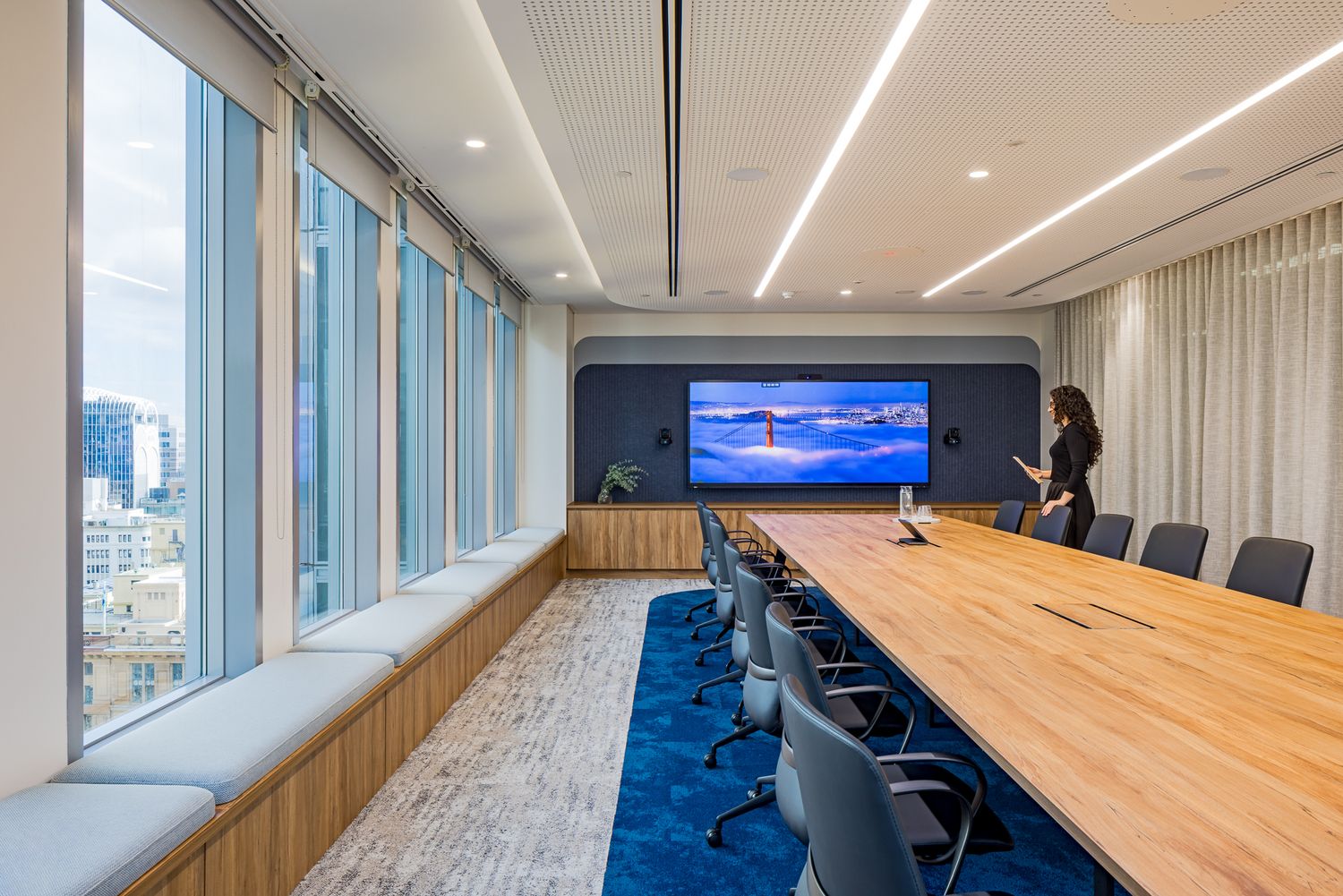 Large boardroom with a long timber table, gray chairs, a wall-mounted video screen and floor-to-ceiling windows with city views.