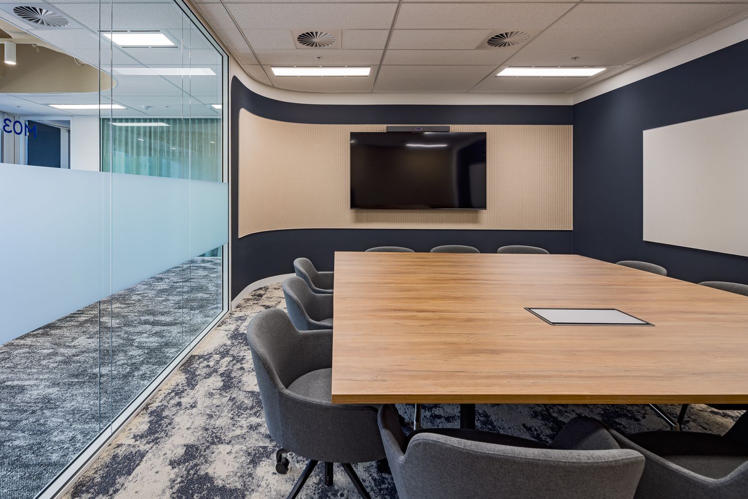 Empty modern meeting room with a large wood table, gray chairs, wall-mounted display and whiteboard beside a frosted glass wall.
