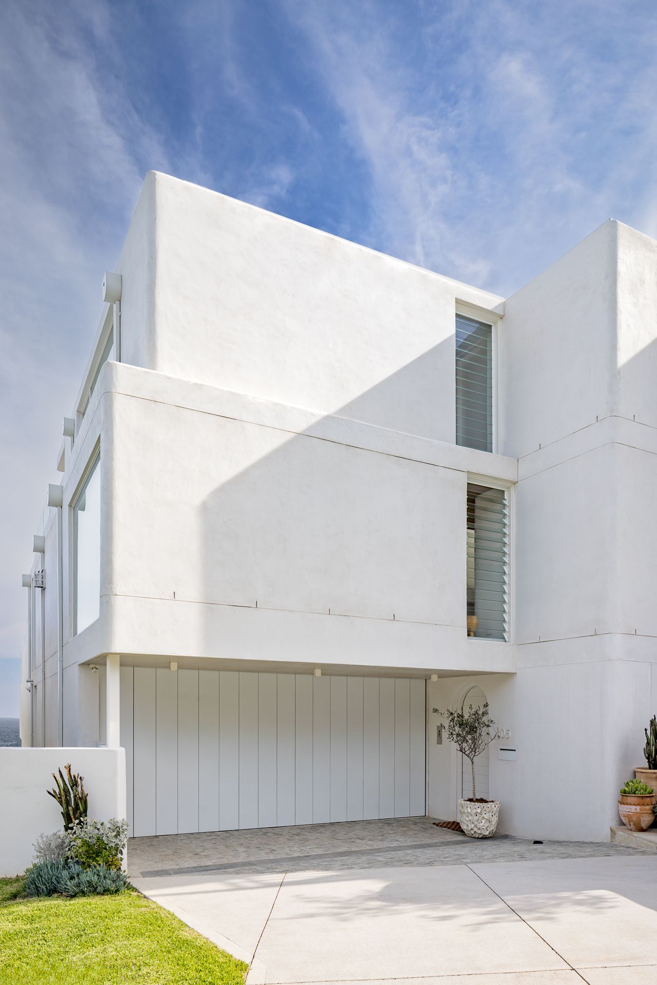 Modern white cubist coastal house exterior with louvered windows, a vertical-panel garage door and potted plants.