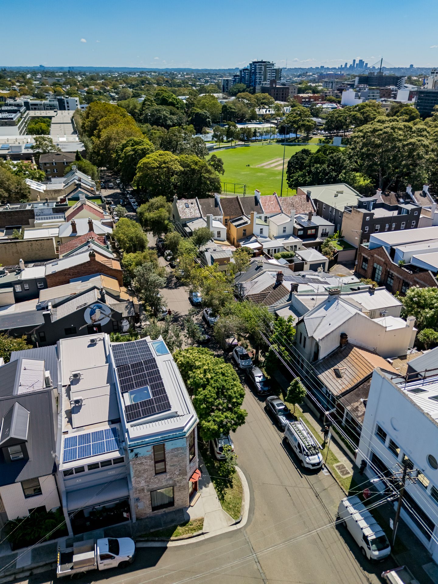 Aerial view of a leafy inner-city neighbourhood with solar-panelled rooftops, terrace houses, a green sports field, and a distant skyline.